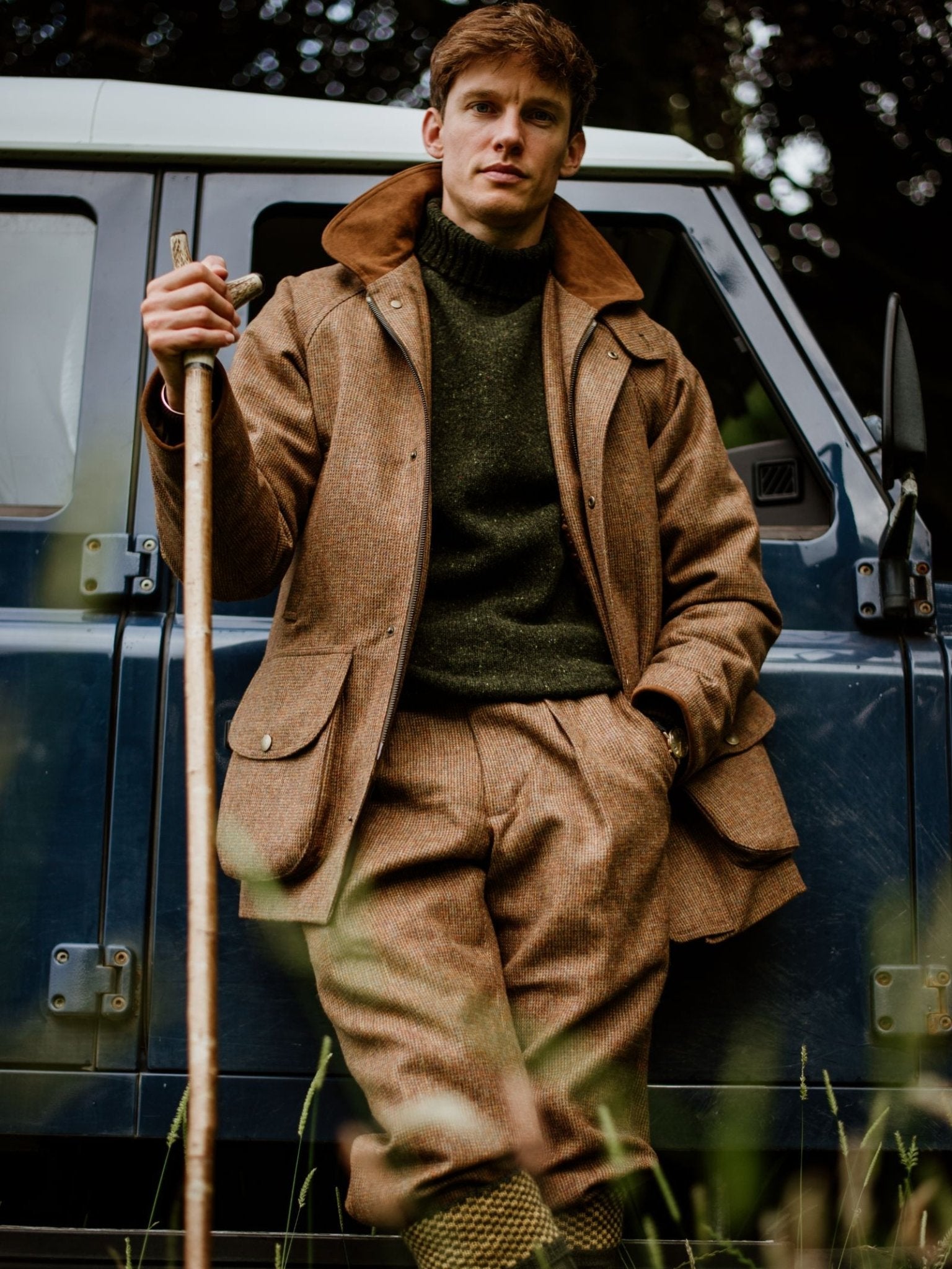 A man in a Campbell's of Beauly Tweed Field Coat, green sweater, and knee-high socks leans on a blue off-road vehicle with a wooden walking stick, the classic UK-made coat standing out against the tall grass and trees behind him.