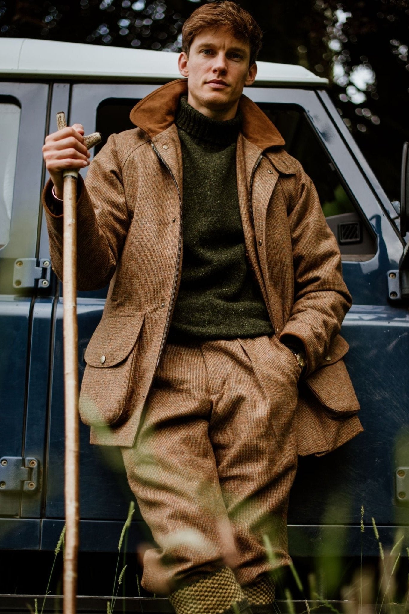 A man in country attire—Campbell's of Beauly Tweed Breeks, a matching Scottish tweed jacket, and a green sweater—leans against a blue vehicle, holding a wooden walking stick amid greenery.