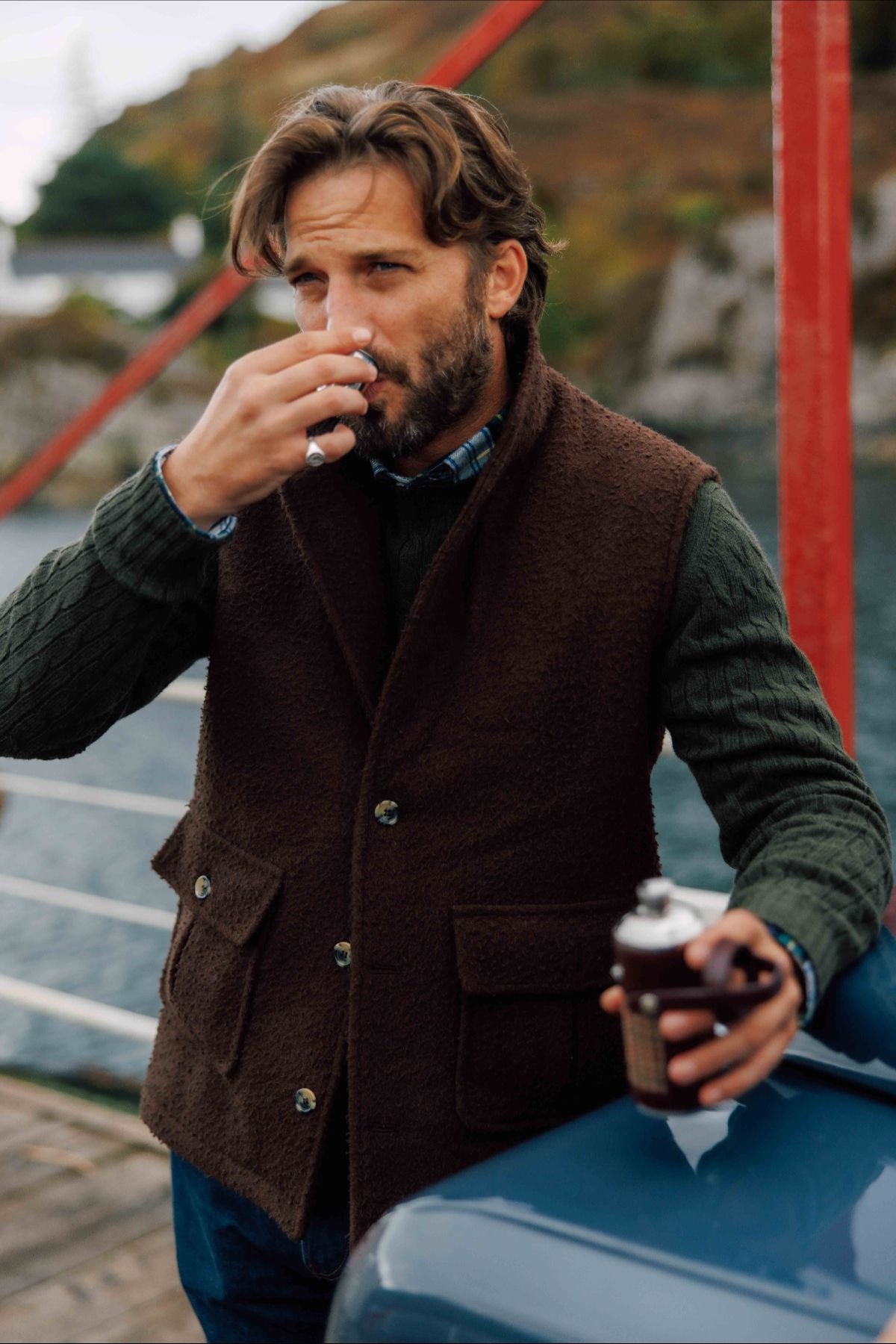 A man with wavy brown hair and a beard, dressed in a green sweater and brown vest, stands outdoors by a body of water, sipping from a small cup and holding a thermos.