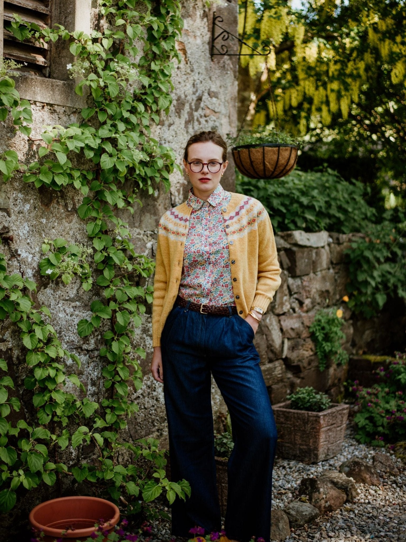A woman wearing glasses, a Campbell's of Beauly Shetland Fairisle Cardigan, a floral shirt, and high-waisted blue pants stands by a stone wall in a rustic garden, embodying classic Scottish knitwear style.