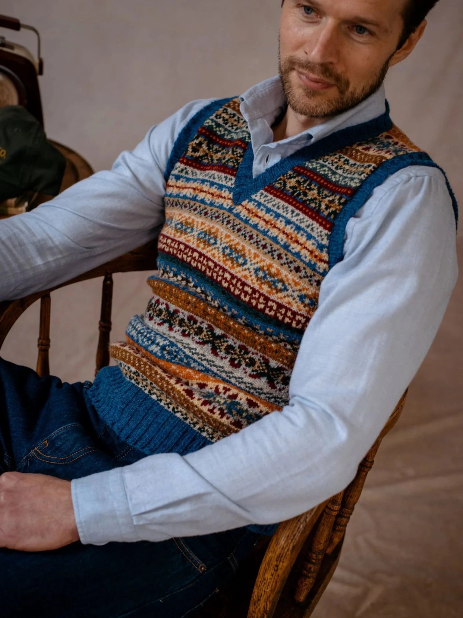 A man sits on a wooden chair wearing a light blue shirt and the Campbell’s of Beauly Fairisle Slipover—a sleeveless layer crafted from Shetland wool, featuring blue, gold, red, and white designs. He has short hair and a trimmed beard, gazing slightly off to the side.