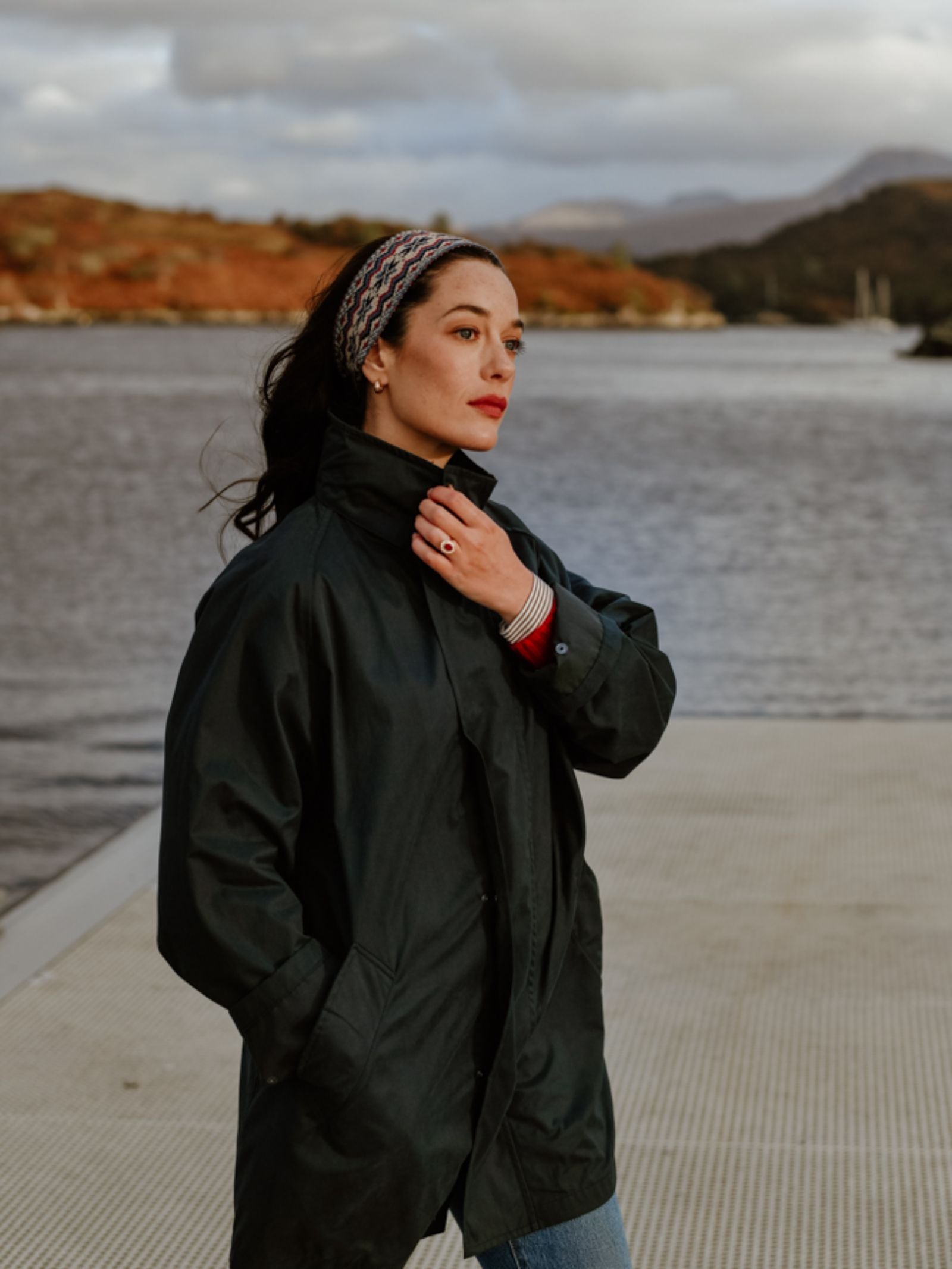 A woman with dark hair and red lipstick stands on a dock by the water, wearing a dark coat and the Campbell’s of Beauly Fairisle Headband, crafted from 100% wool; Scottish Borders hills and trees rise in the background.