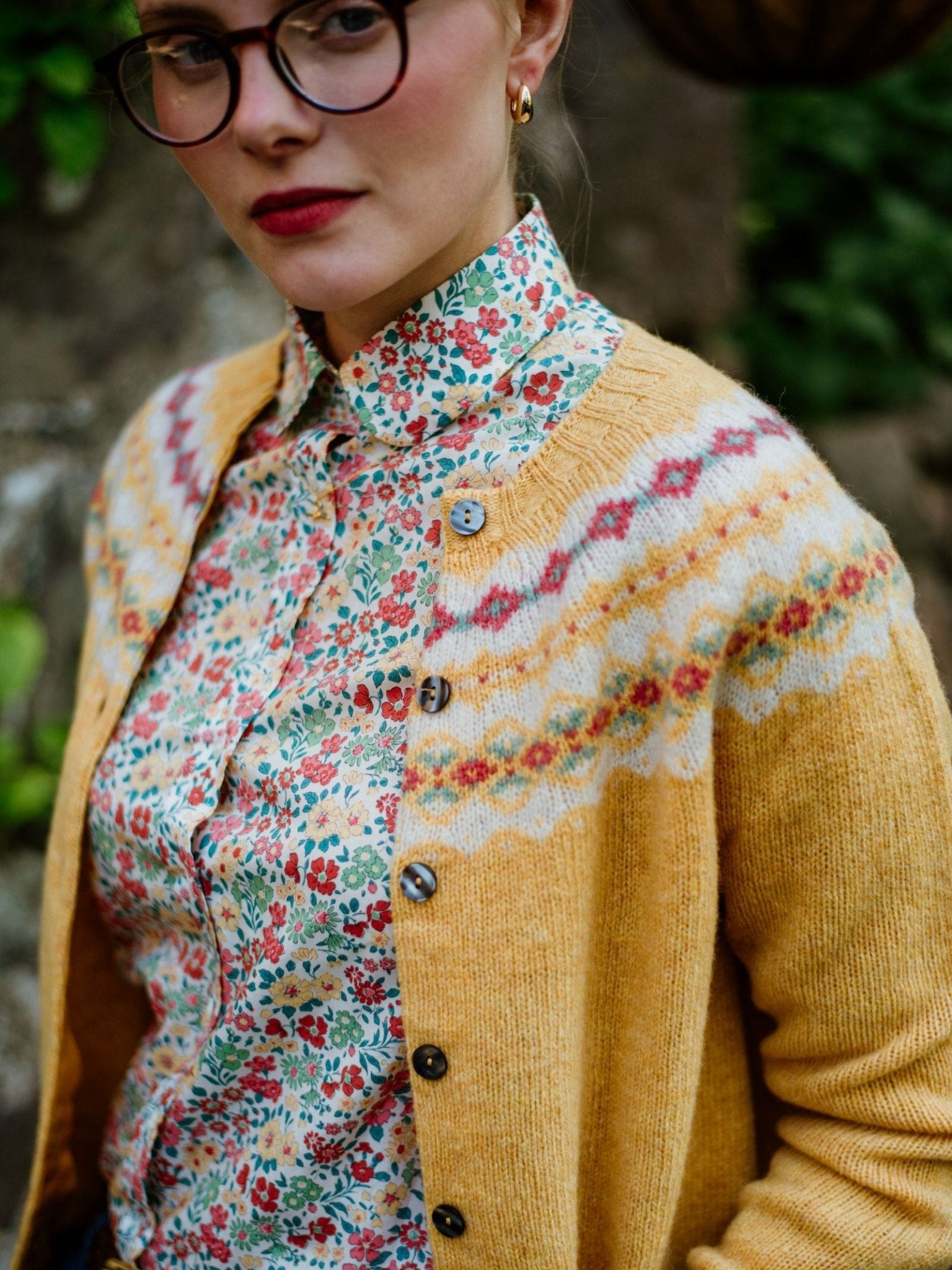 A person wearing round glasses, a yellow patterned cardigan, and a Campbells of Beauly Liberty Print Cotton Shirt stands outdoors. The upper body and face are shown, highlighting thoughtful expression and vintage-inspired style.