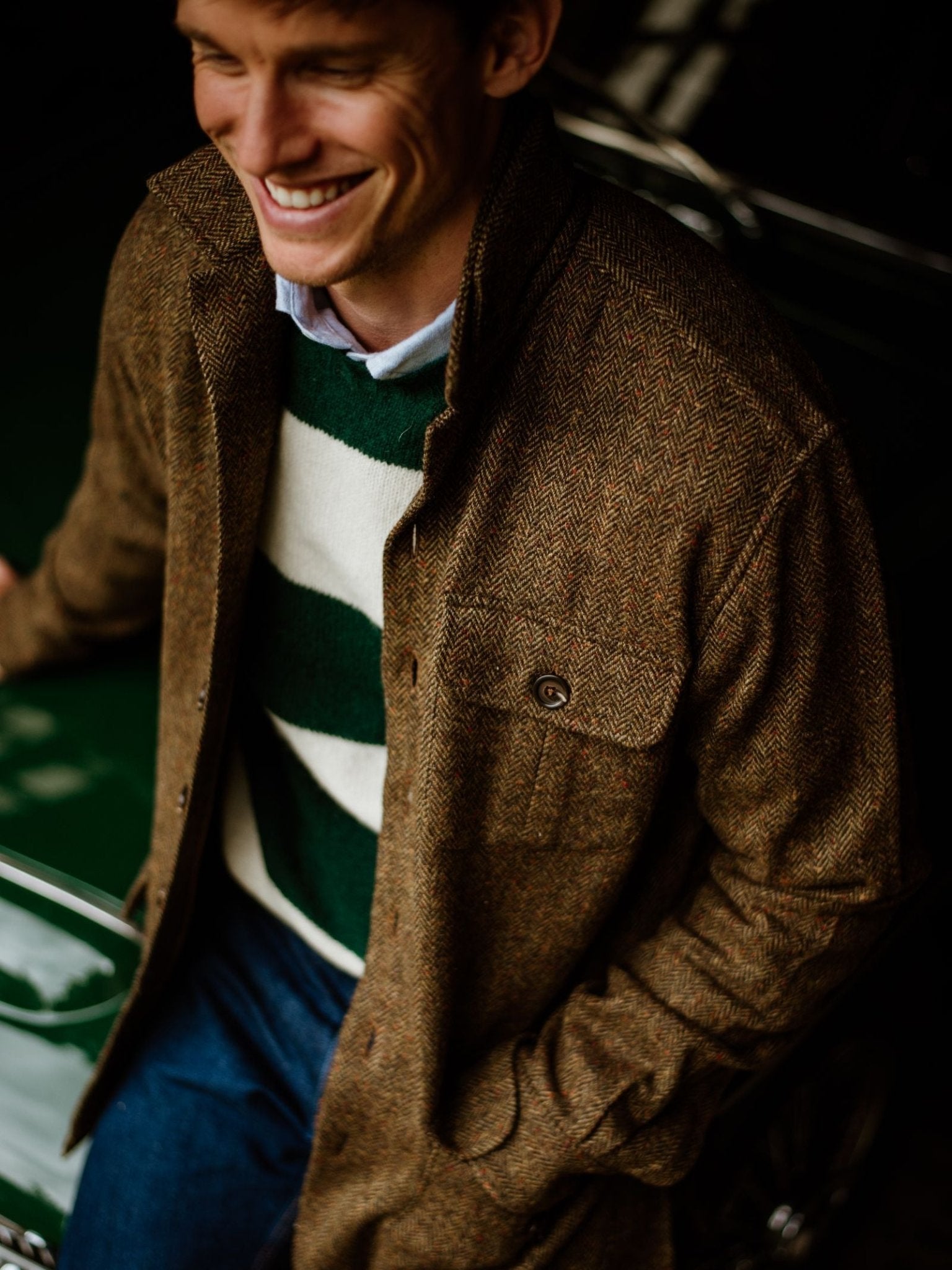 A man in the Campbell's of Beauly Casual Tweed Shacket, layered over a green and white striped sweater, stands by a dark green car with one hand in his pocket, smiling.