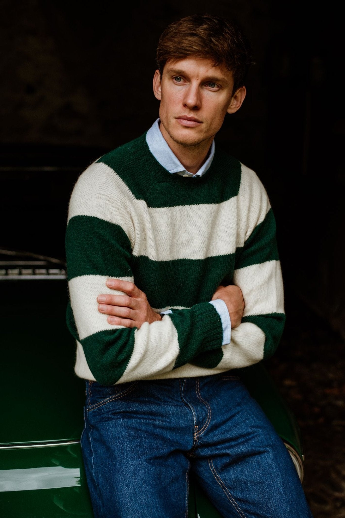 A man in a green and white Auld Stock Shetland Stripe Crew Jumper by Campbell's of Beauly, layered over a light blue collared shirt and blue jeans, sits with arms crossed against a dark background, gazing to the side.