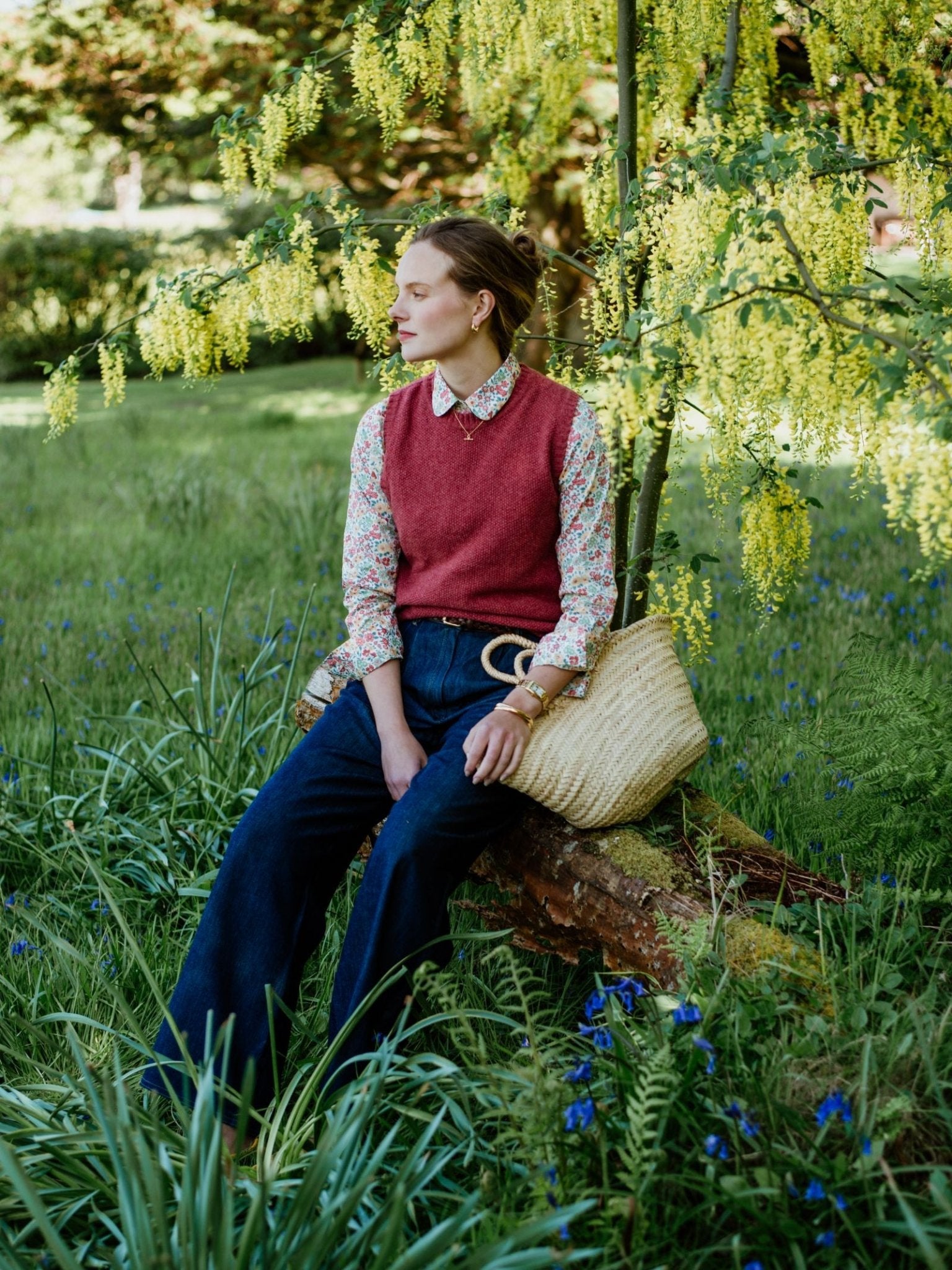 A woman in a Campbells of Beauly Moss Stitch Crew Tank, floral shirt, and wide-leg jeans sits on a log in a grassy garden with a straw bag, surrounded by green foliage and yellow flowers—a perfect pick for versatile layering.
