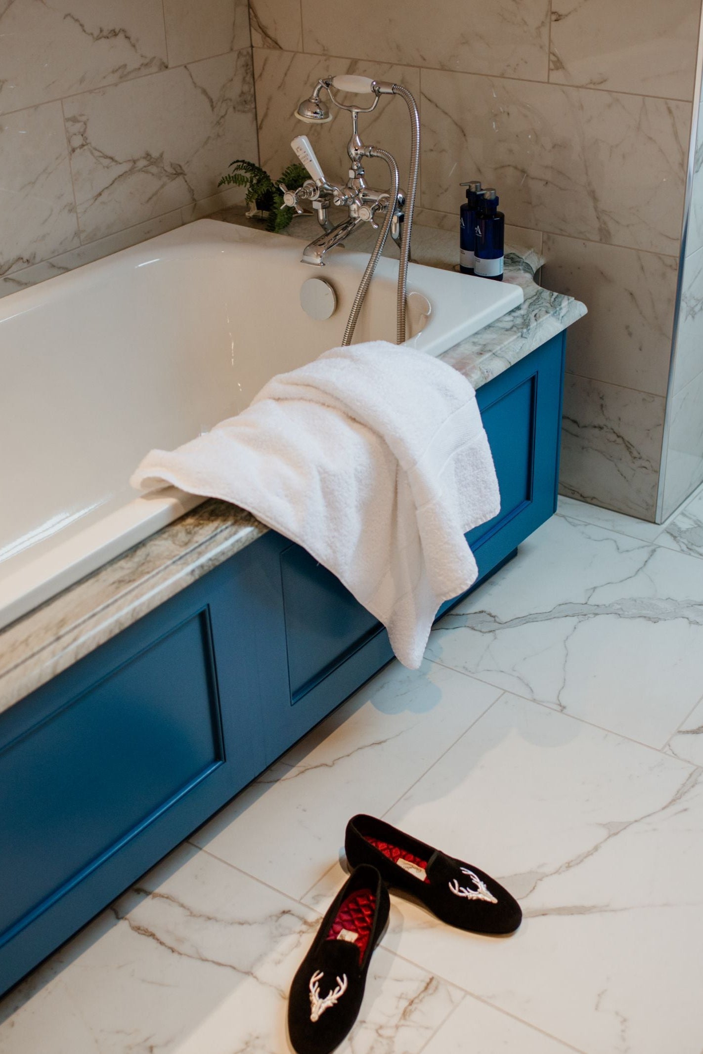 A white towel hangs over a bathtub with blue paneling and marble trim. On the floor are white tiles and a pair of Campbell's of Beauly Stag Velvet Slippers with an embroidered stag motif. A plant and bottles sit on the tub ledge.