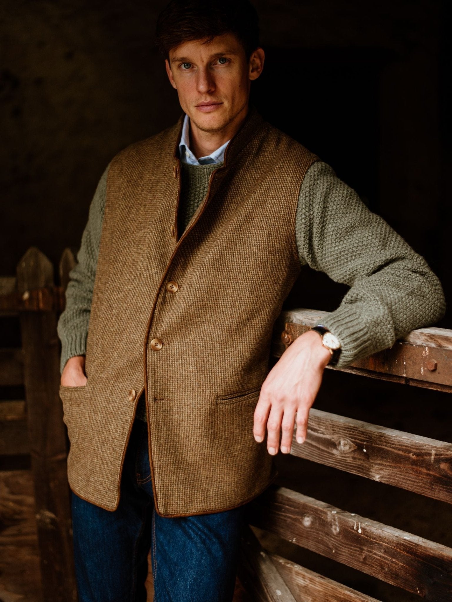 A man wears the Campbell's of Beauly Tweed Nehru Gilet over a green sweater and blue jeans, leaning casually against a wooden fence in a rustic, dimly lit setting. The gilet is crafted from classic British tweed.