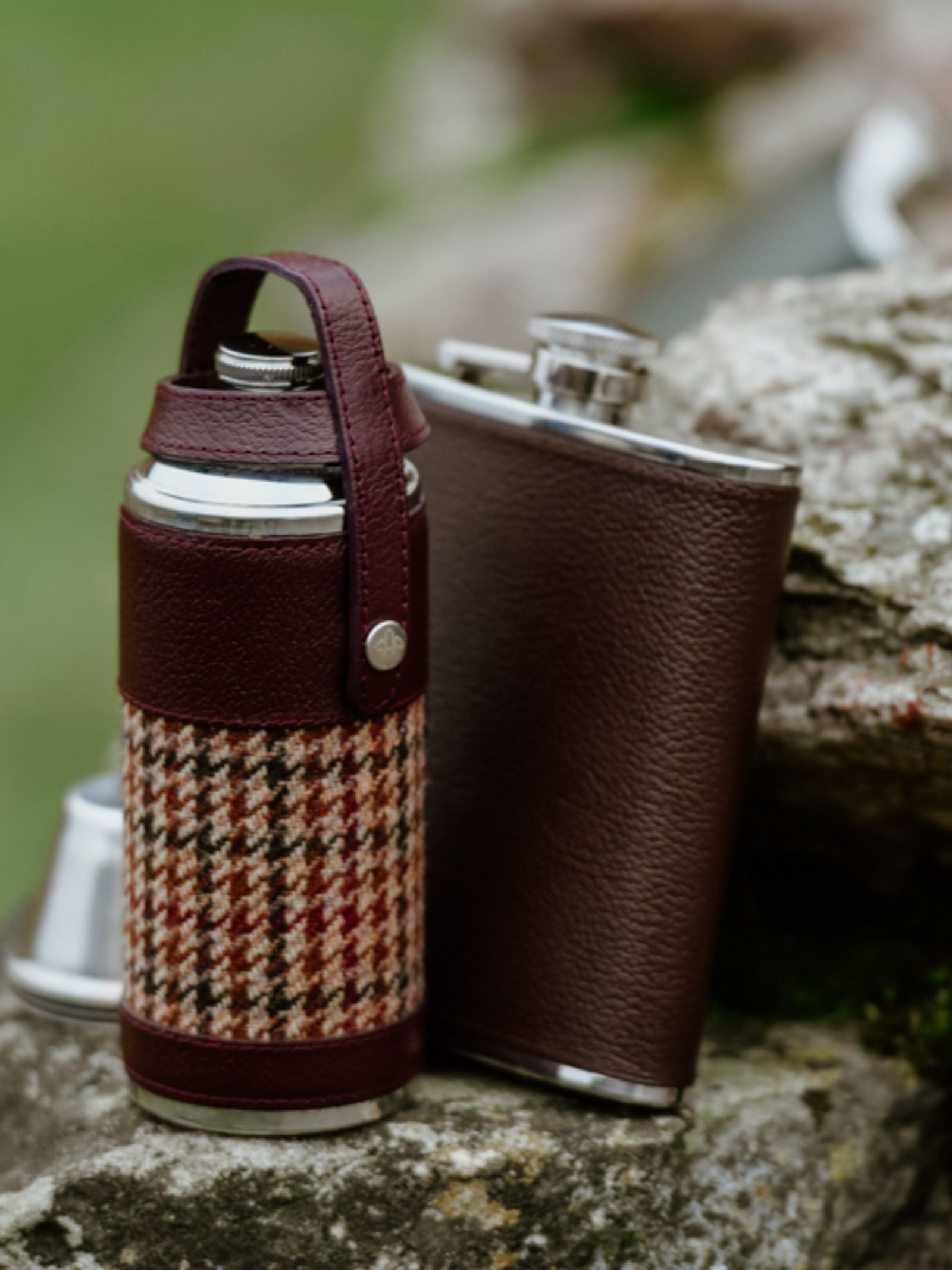 Two 8oz Bordeaux Hunter Flasks by Campbell's of Beauly x Ettinger rest outdoors; one is wrapped in brown Capra goat leather with houndstooth fabric, the other in plain leather. Both feature British craftsmanship and sturdy metallic caps.