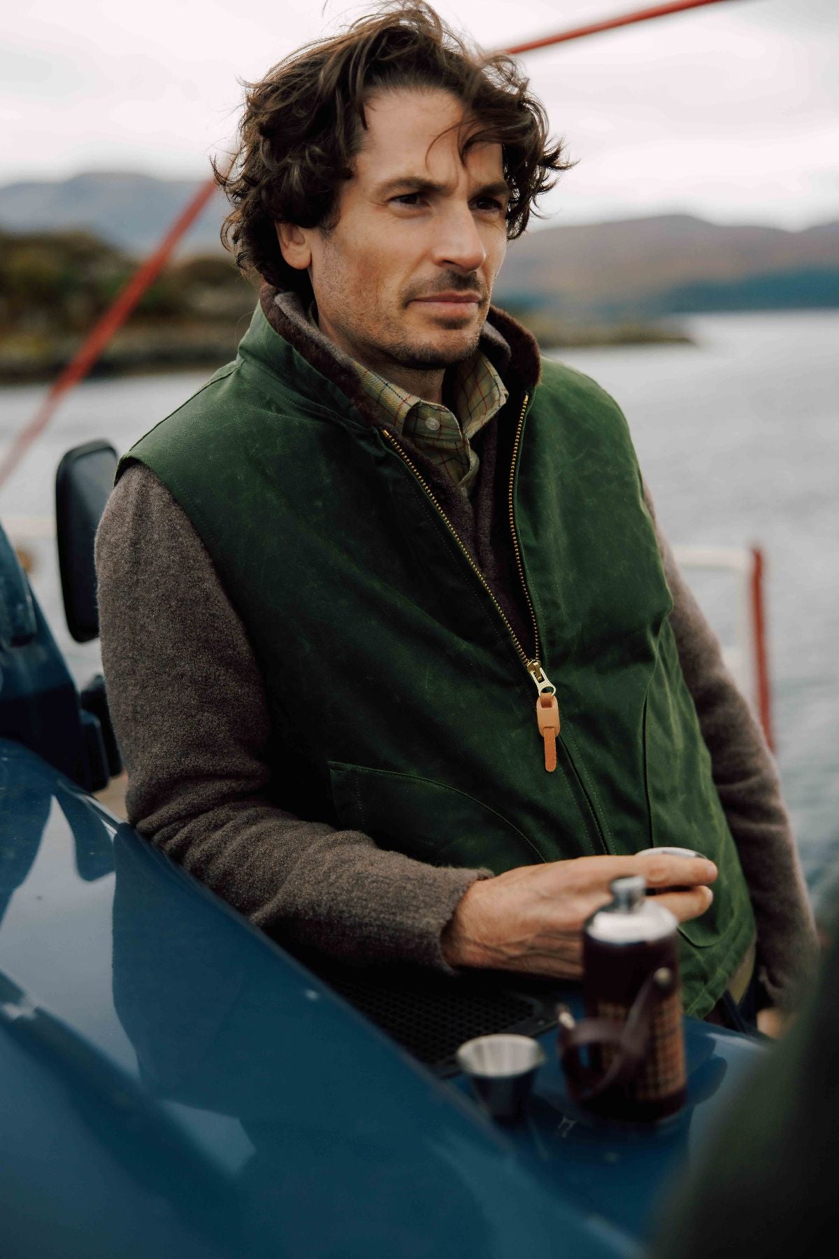 A man with wavy brown hair, wearing a green vest and brown sweater, leans against a blue vehicle by a body of water, holding a drink with a thermos and cup on the hood.