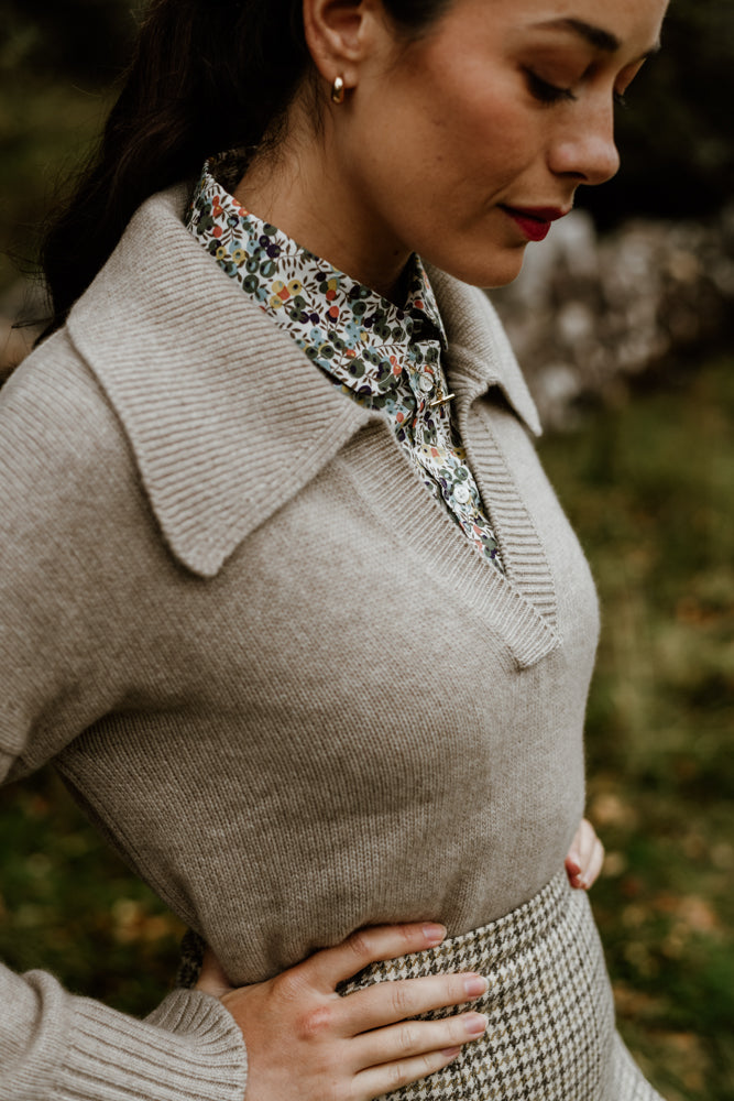 A woman stands outdoors with her hand on her hip, wearing a beige knit sweater over a floral collared shirt and a houndstooth skirt. Her hair is tied back, and she has red lipstick and small hoop earrings.
