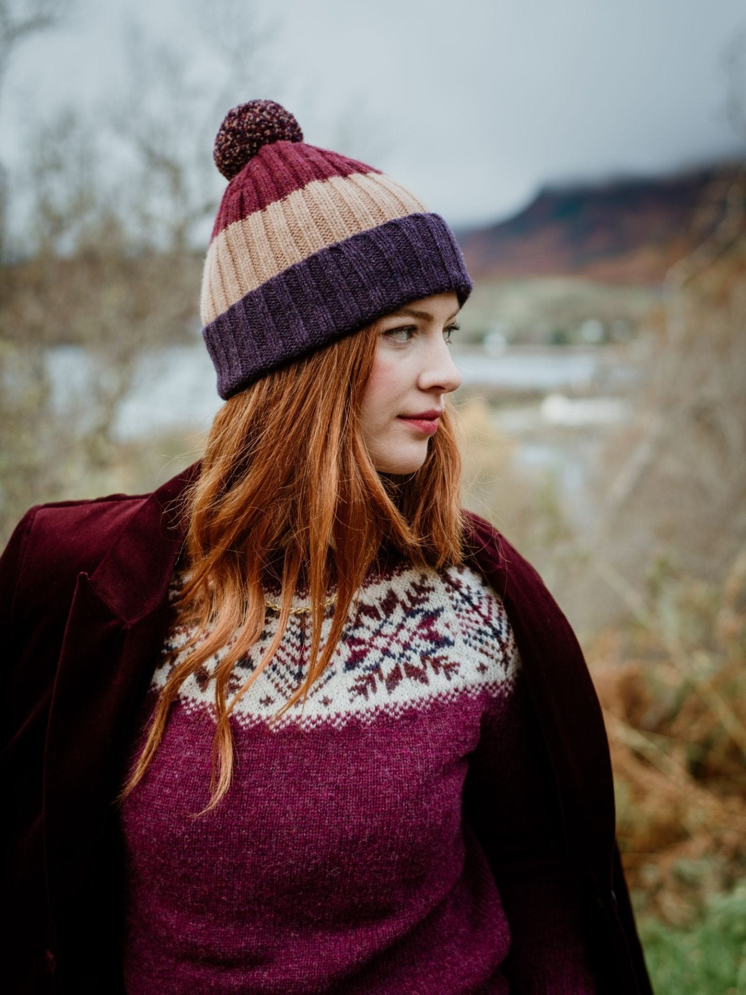 A woman with long red hair wears a Campbell's of Beauly Auld Stock Lambswool Stripe Hat with Pom-Pom, a maroon coat, and a patterned sweater while standing outdoors among blurred trees and distant mountains.