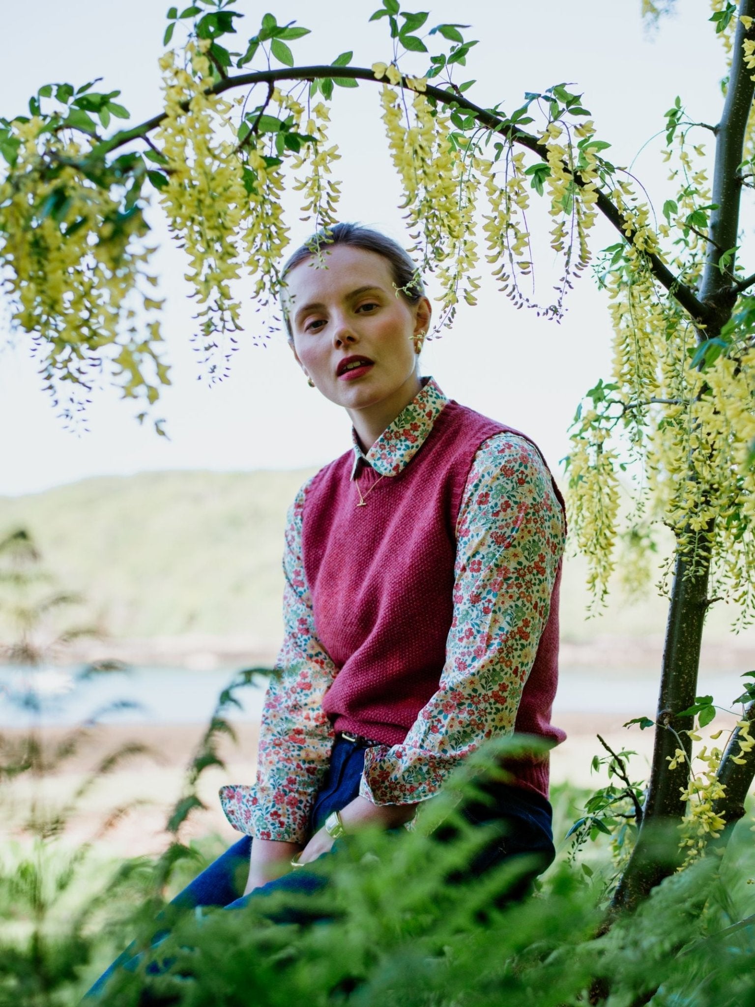 A person in the Auld Stock Moss Stitch Crew Tank by Campbell's of Beauly and a floral shirt sits outdoors beneath yellow flowers, surrounded by greenery, with blurred hills and water in the background.
