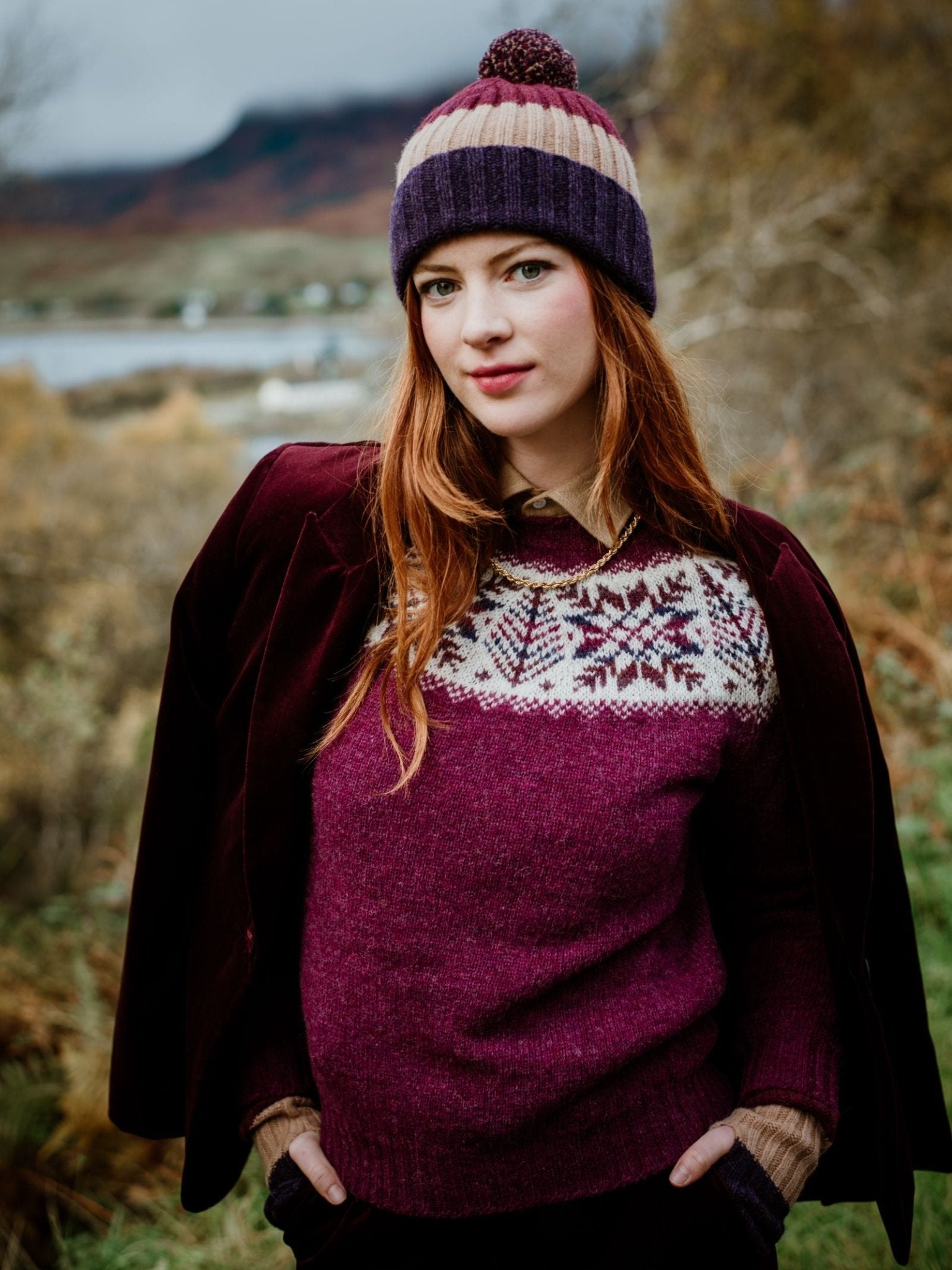 A woman with long red hair stands in the Scottish Borders countryside, wearing Campbell's of Beauly's Auld Stock Lambswool Stripe Hat with Pom-Pom, along with a purple and cream sweater and jacket. Autumn colors fill the background.