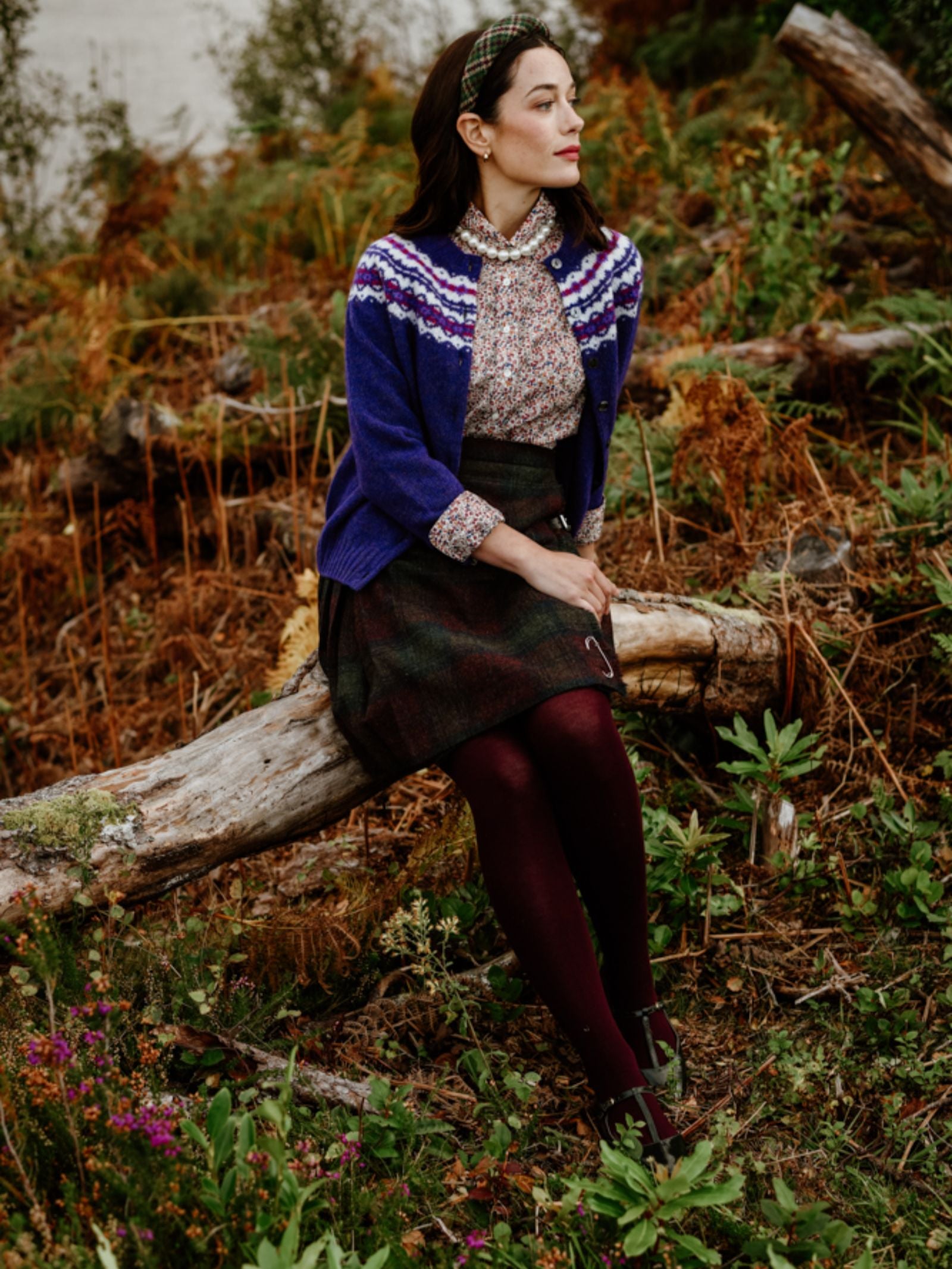 A woman wearing a blue patterned cardigan, floral blouse, plaid skirt, and burgundy tights sits on a log in a forest setting, surrounded by greenery and autumnal plants. She looks pensively to the side.