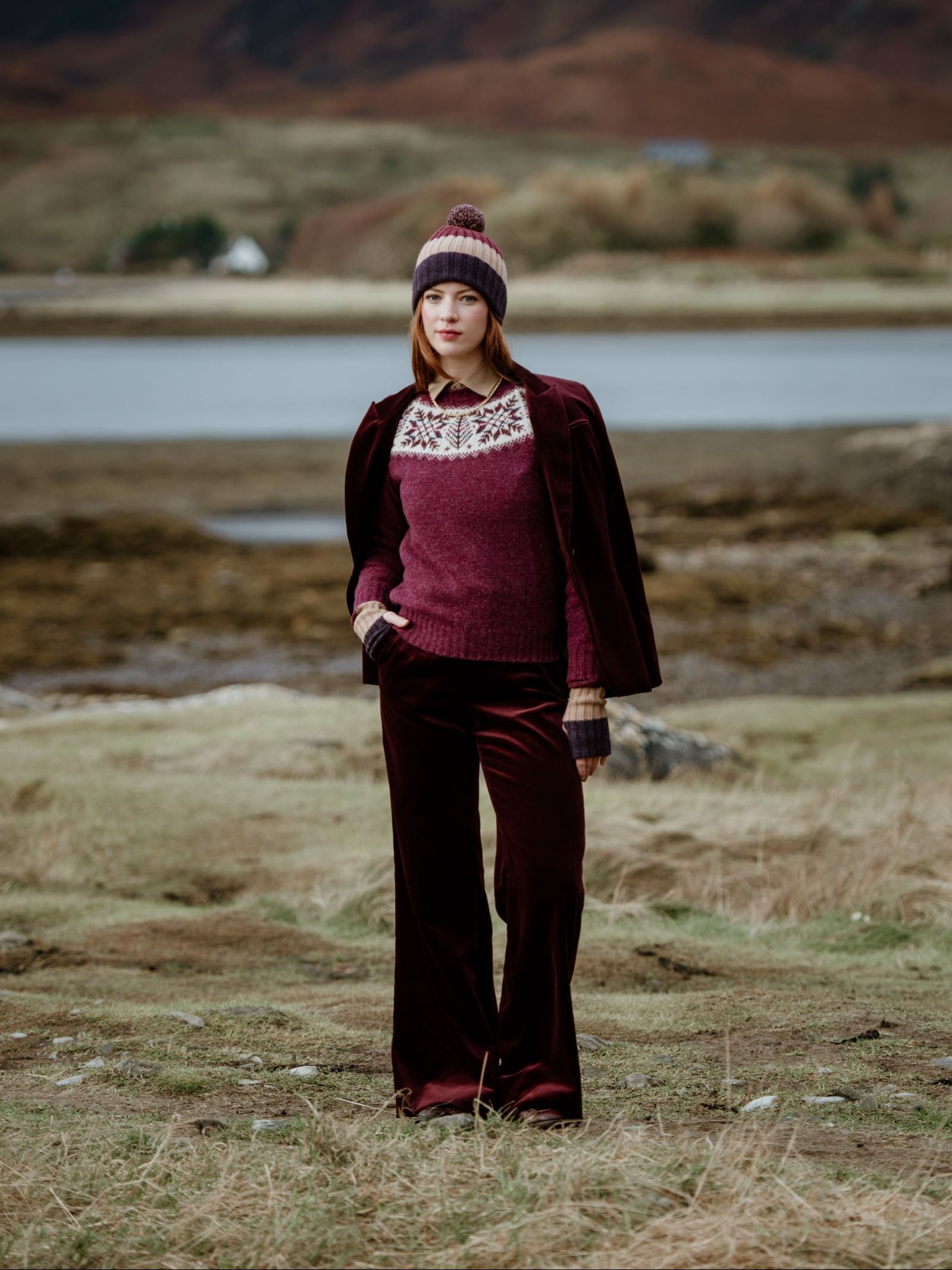 A woman stands outdoors near water and hills in the Scottish Borders, wearing the Campbell's of Beauly Auld Stock Lambswool Stripe Hat with Pom-Pom, a patterned sweater, a jacket over her shoulders, and matching velvet pants, hands in her pockets.