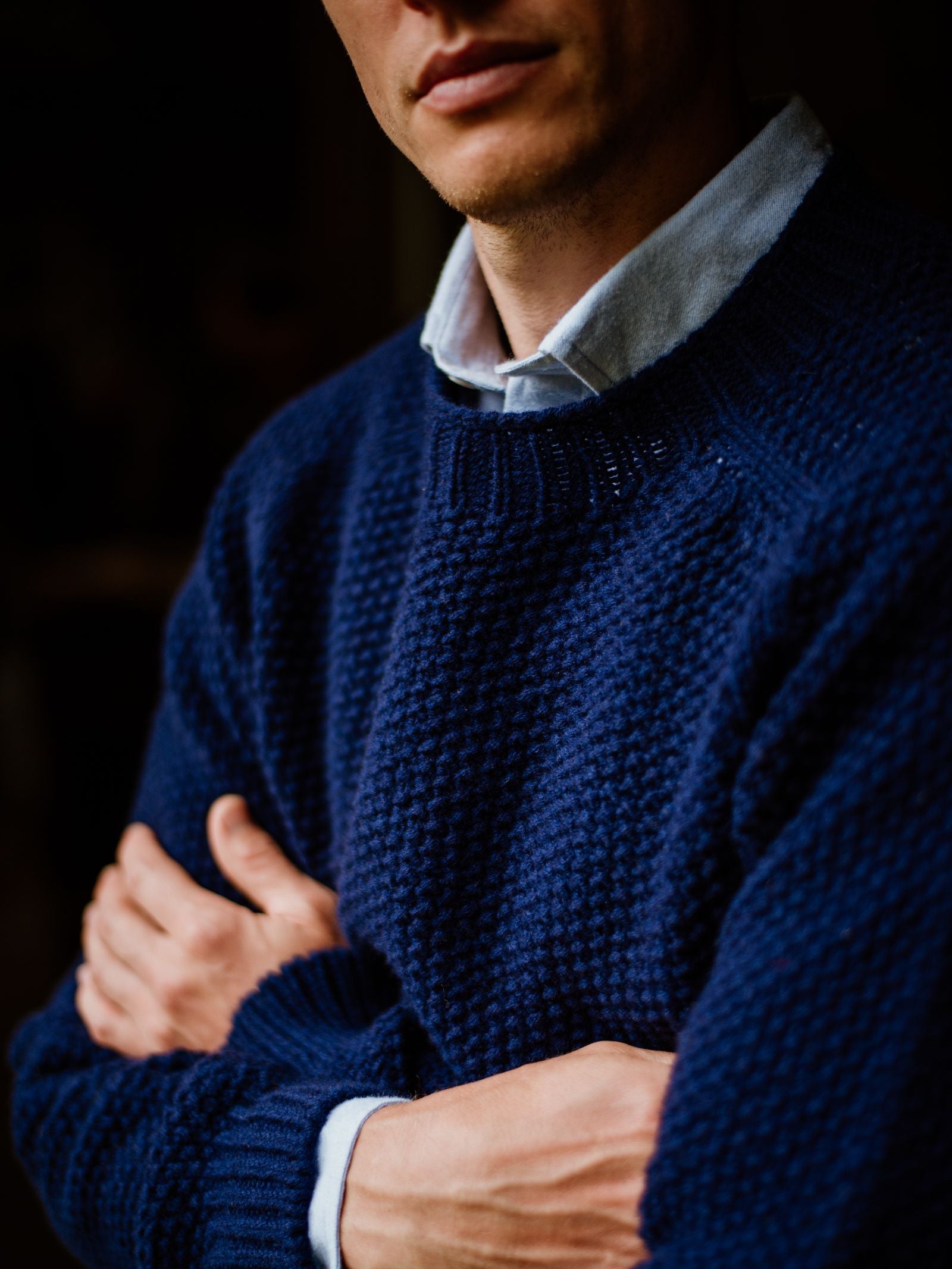 A person wearing a textured navy blue sweater and a light blue collared shirt stands with arms crossed, shown from the shoulders down, with their face partially visible.