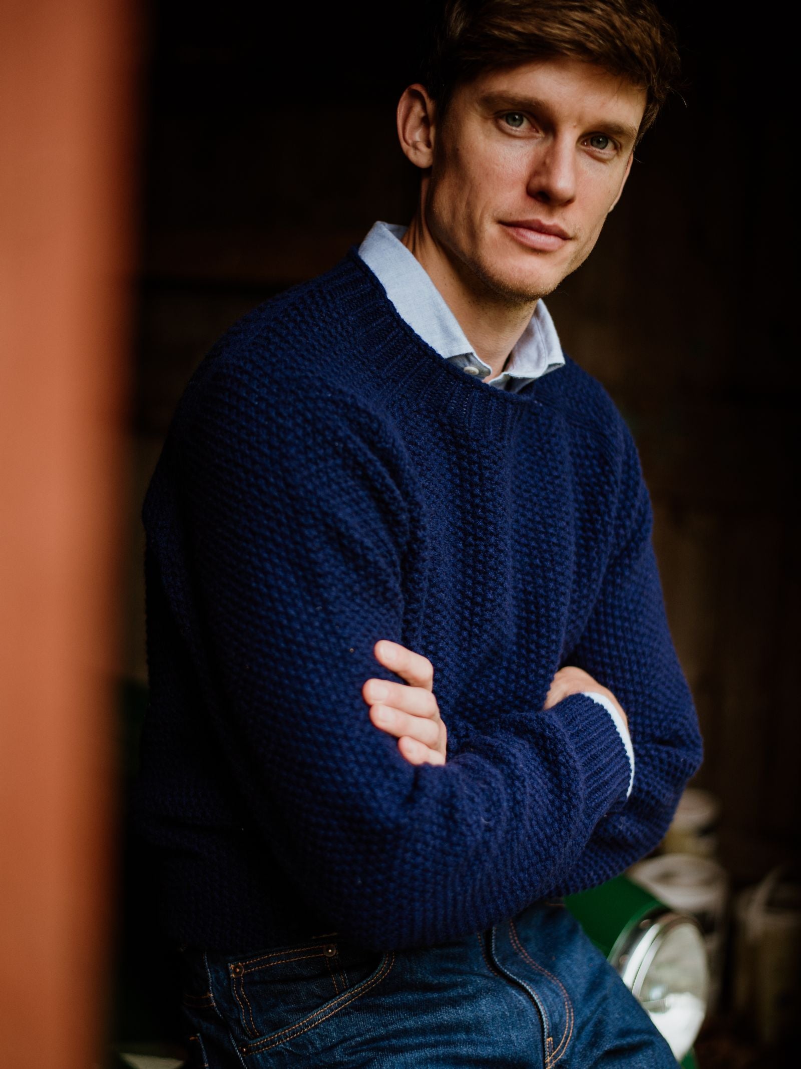 A man with light brown hair wears a Campbell’s of Beauly Auld Stock Moss Stitch Jumper, knitted in Scotland, over a light blue collared shirt and jeans. He stands indoors with his arms crossed, looking at the camera.
