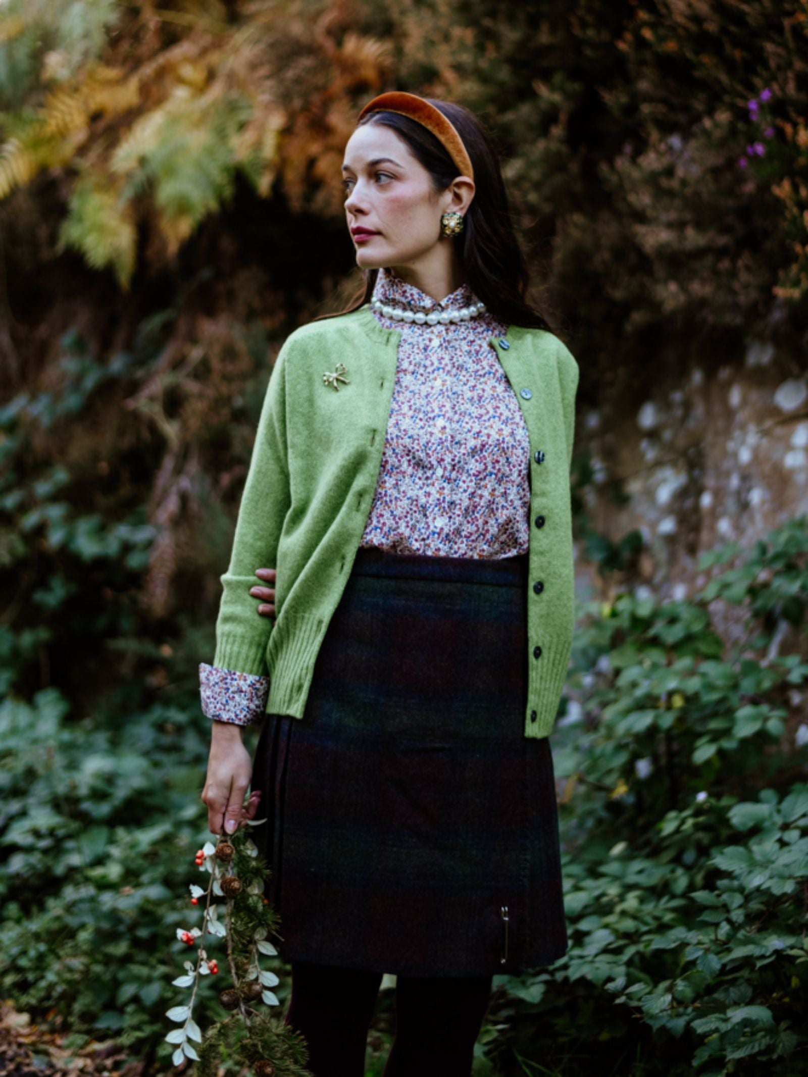 A woman outdoors wears the Campbell’s of Beauly Geelong Lambswool Crew Cardigan in Fern, paired with a floral blouse, dark plaid skirt, and brown headband. She holds a small bouquet amid greenery and autumn foliage.