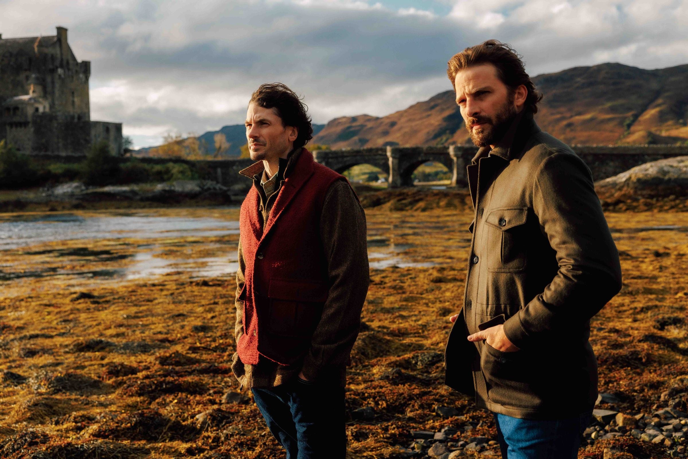 Two men stand on a rocky shoreline with a castle and stone bridge in the background, surrounded by hills under a cloudy sky. Both wear jackets and look thoughtfully into the distance.
