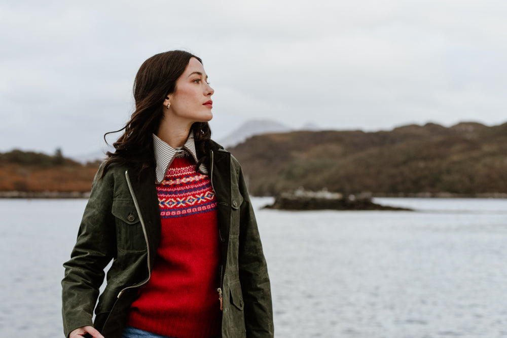 A woman in a green jacket and red patterned sweater stands by a lake, looking to the side. Hills and cloudy sky are in the background, creating a calm, natural setting.