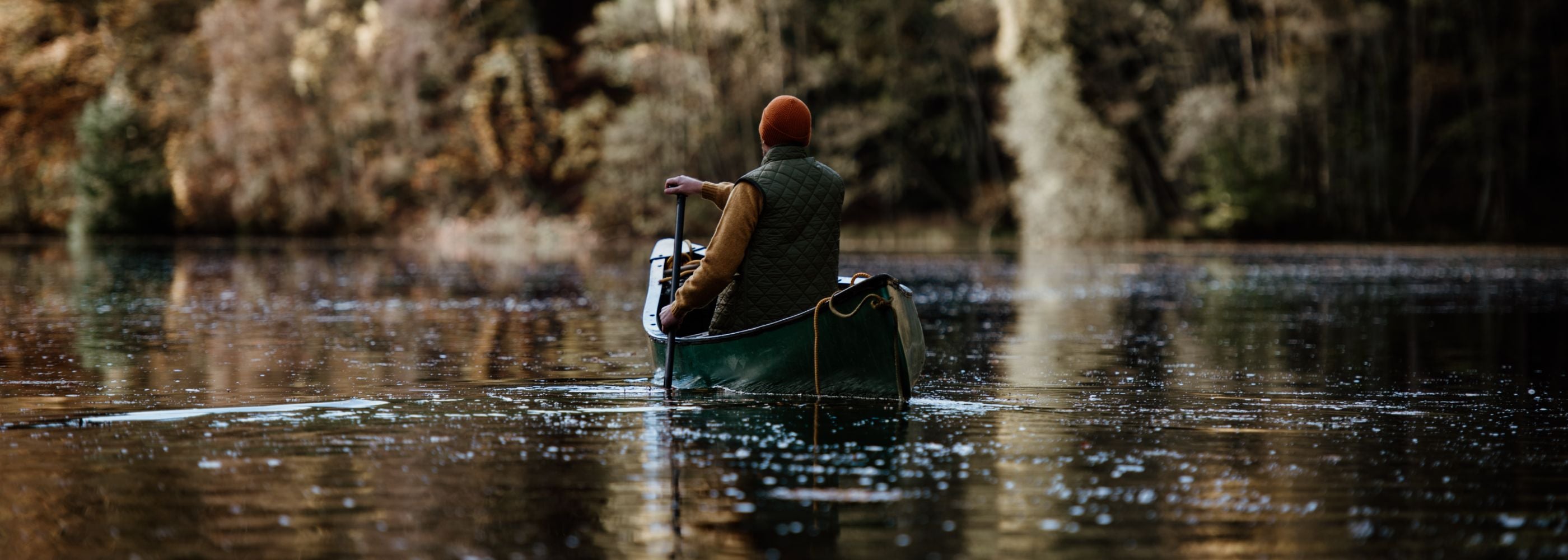 A person wearing an orange beanie and green vest paddles a green canoe on a calm lake, surrounded by autumn trees reflected in the water.