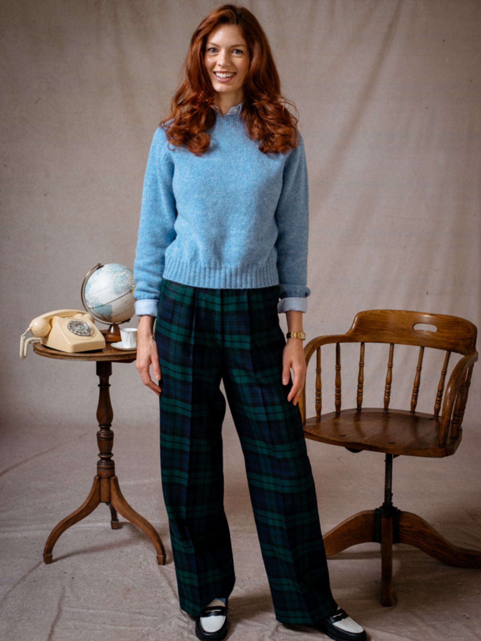A woman with long red hair wears the Campbell's of Beauly Auld Stock Cropped Shetland Jumper in light blue and green plaid pants, smiling indoors by a wooden chair and table with a globe, rotary phone, and pocket watch. Neutral backdrop.