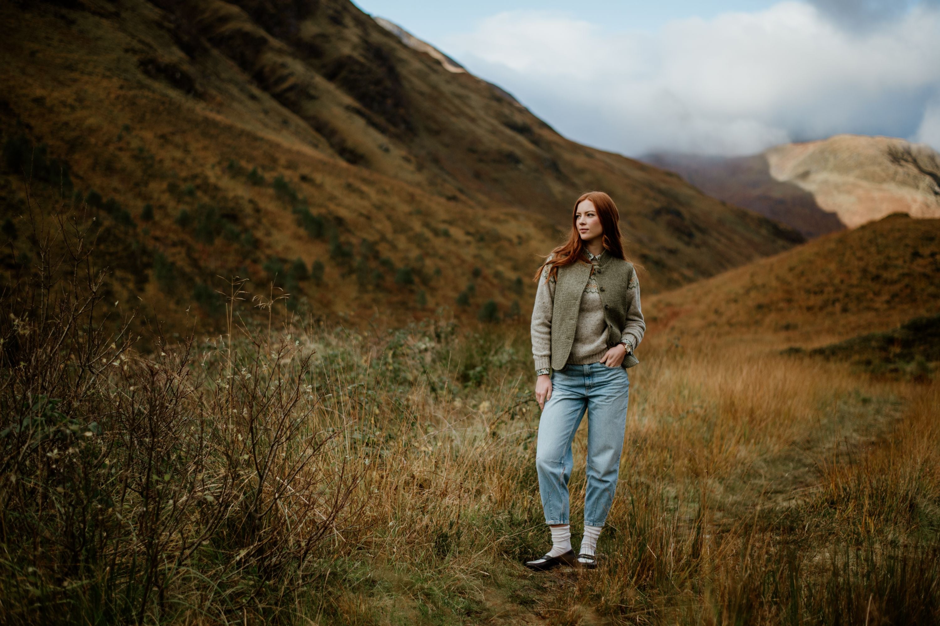 A woman with long red hair, wearing a sweater and jeans, stands on a grassy path surrounded by hills covered in autumn foliage under a partly cloudy sky.