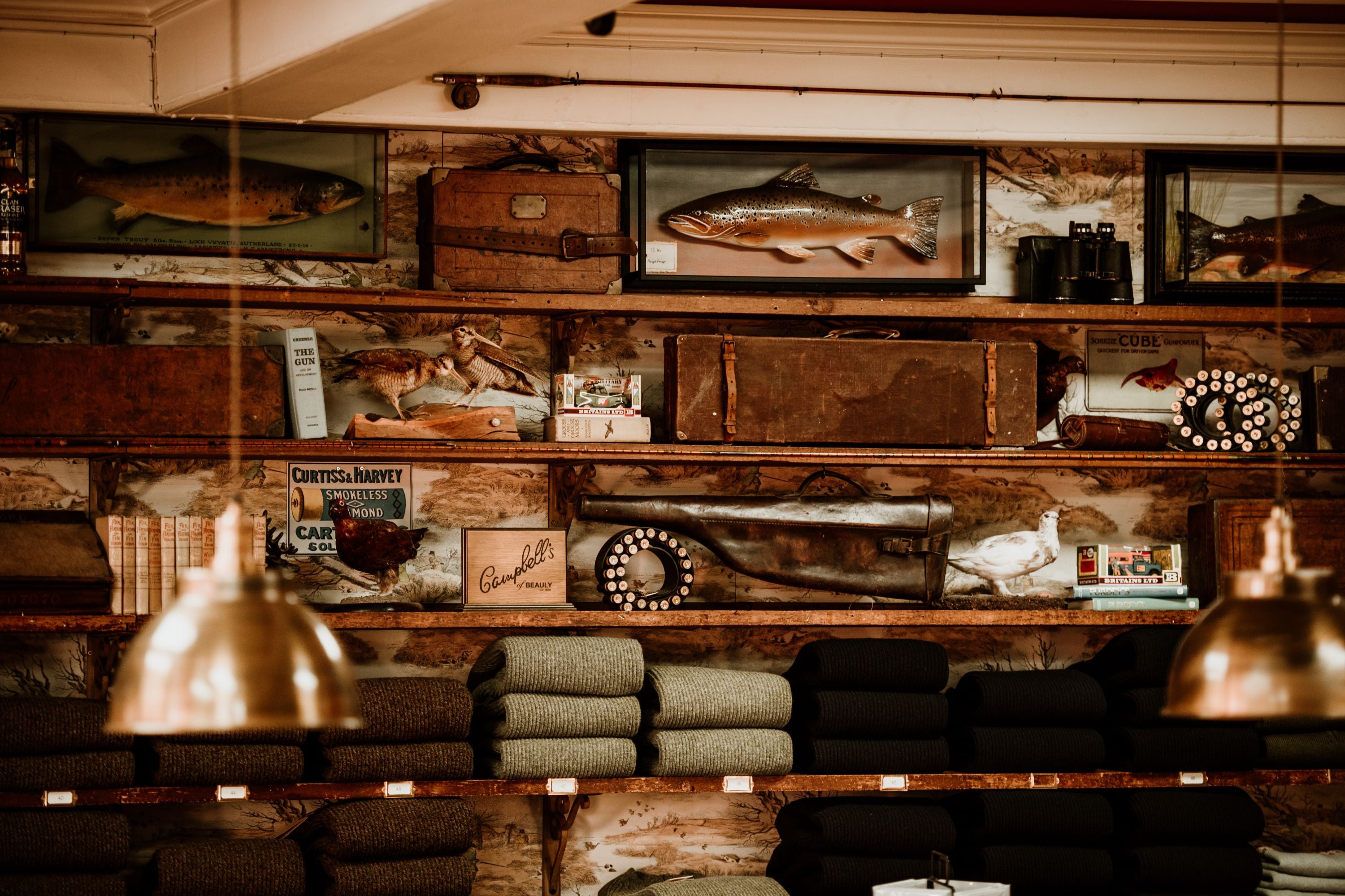 Shelves filled with vintage suitcases, framed fish, books, old cameras, and fishing gear, above neatly stacked folded sweaters, with hanging brass pendant lights in the foreground.