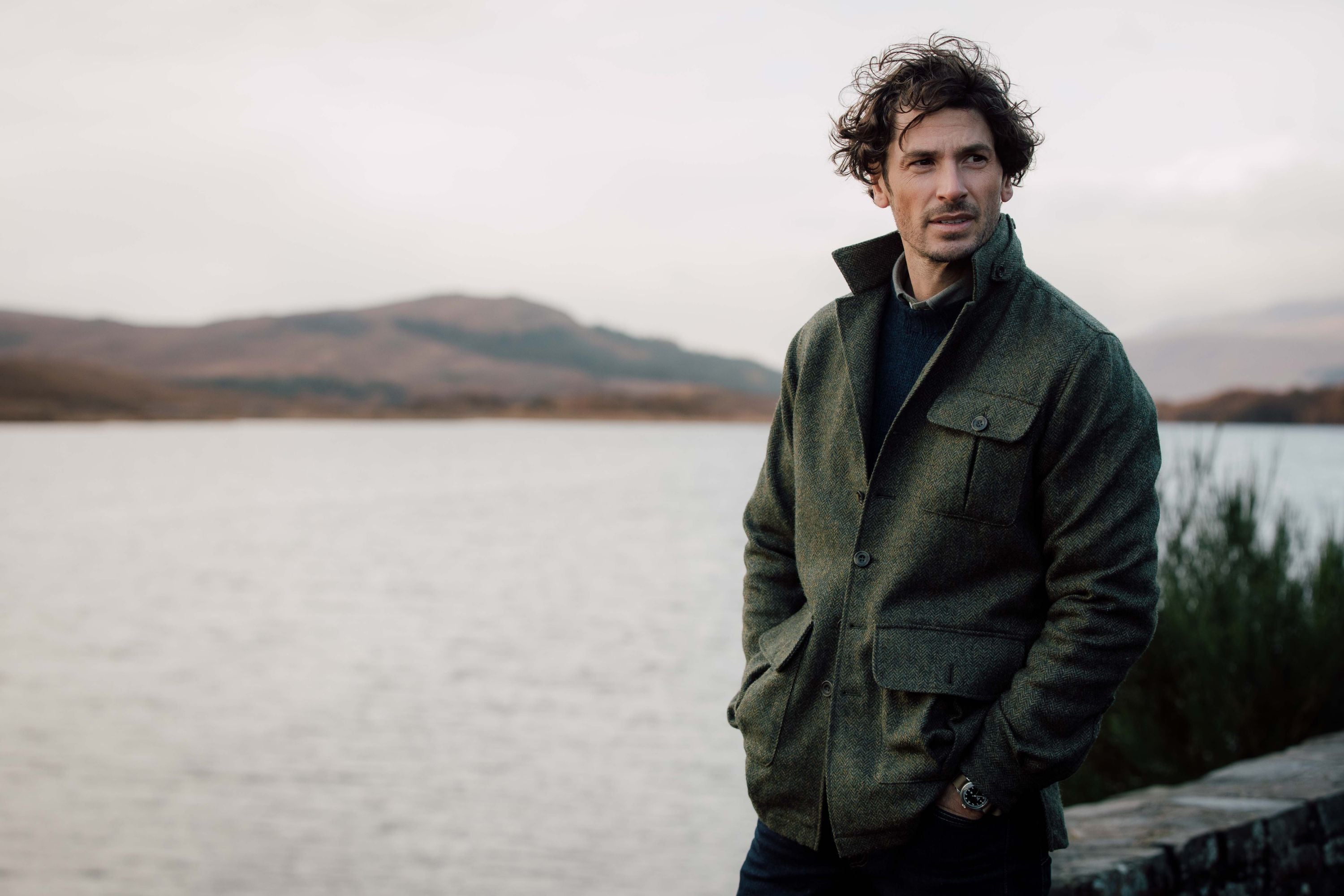 A man with curly hair wearing a green jacket stands outdoors by a lake, with hills and cloudy sky in the background. He has his hands in his pockets and looks to the side, appearing thoughtful.