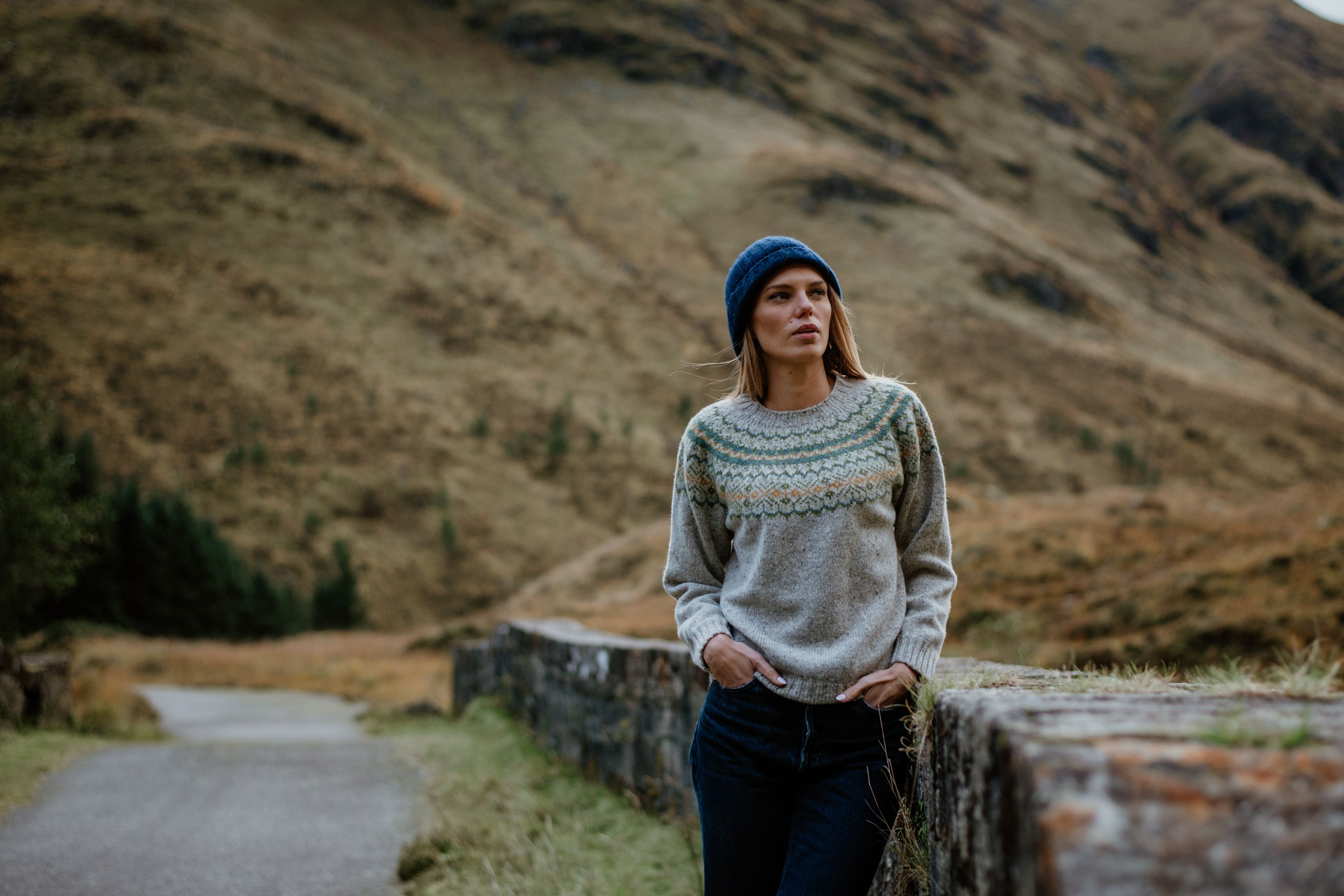 A woman in a blue knit hat and patterned sweater leans against a stone wall on a rural path, with rolling hills and grass in the background under an overcast sky.