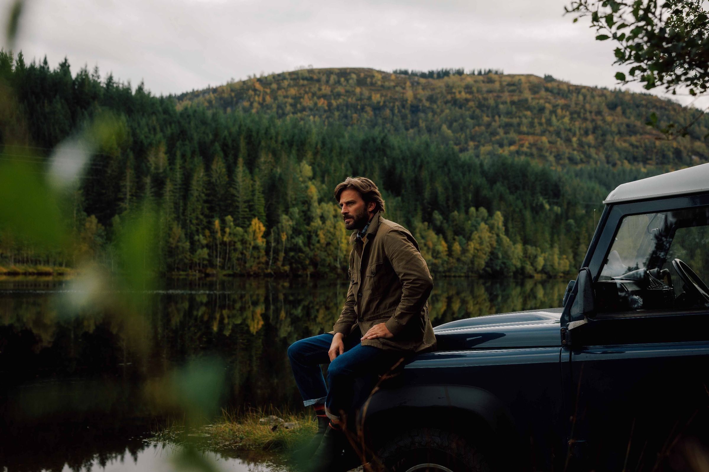 A man sits on the hood of a dark vehicle by a calm lake, surrounded by pine trees and forested hills under a cloudy sky. Greenery and reflections are visible in the peaceful, natural setting.