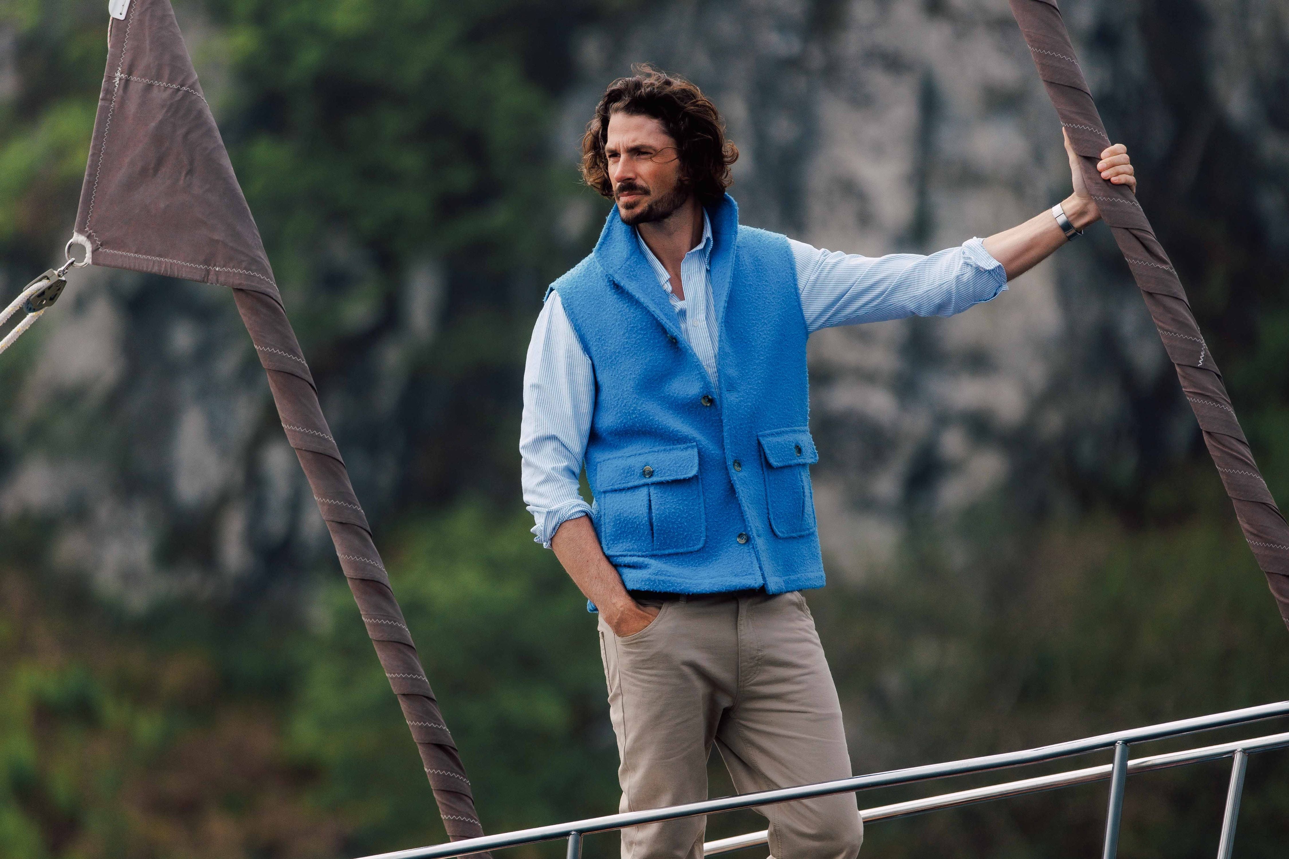 A man with wavy hair, wearing a light blue shirt, blue vest, and beige pants, stands on a boat, holding a rope and looking into the distance with trees and rocky cliffs in the background.