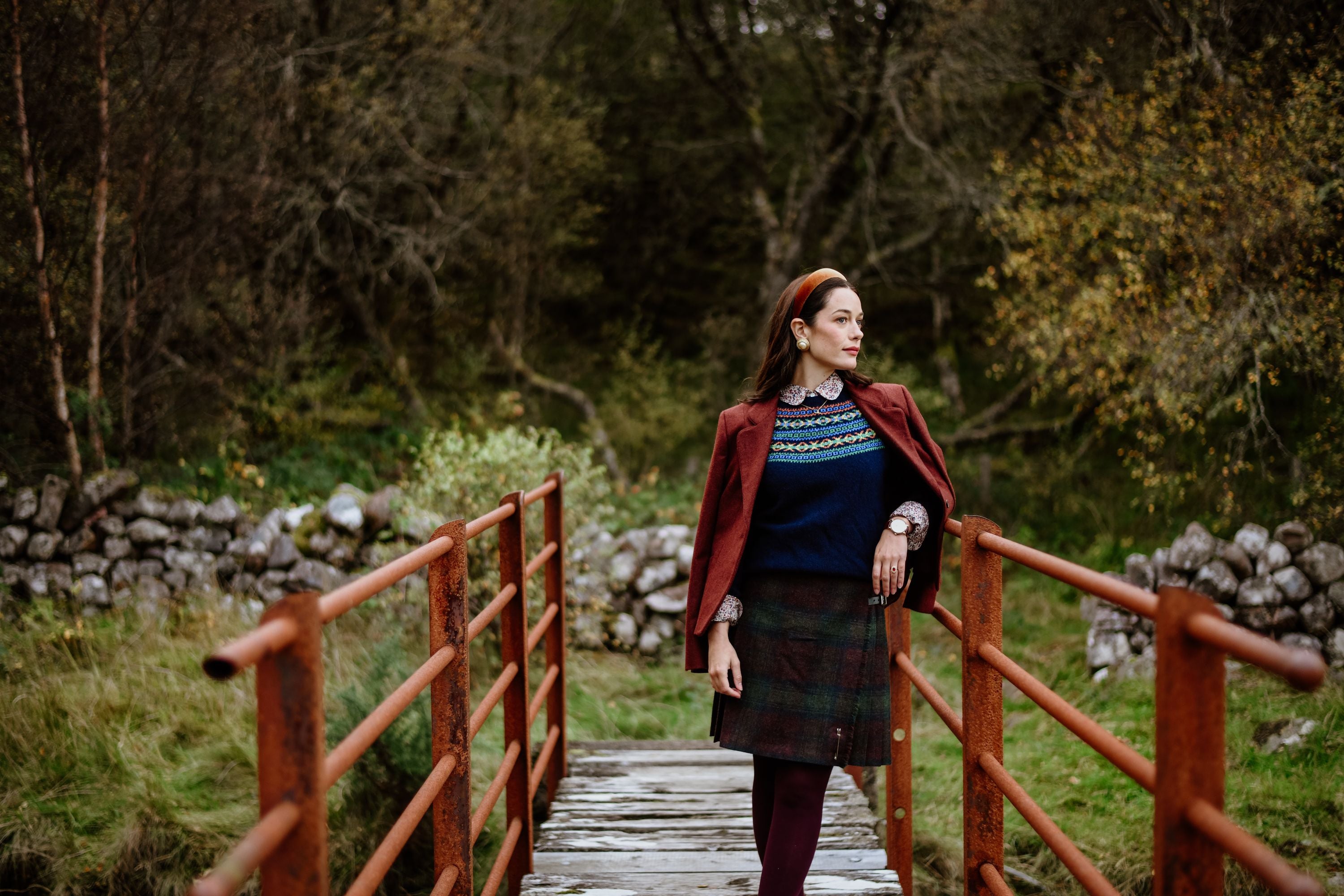 A woman in a plaid skirt, navy sweater, and maroon jacket stands on a rustic red bridge in a natural setting with trees and stone walls in the background.