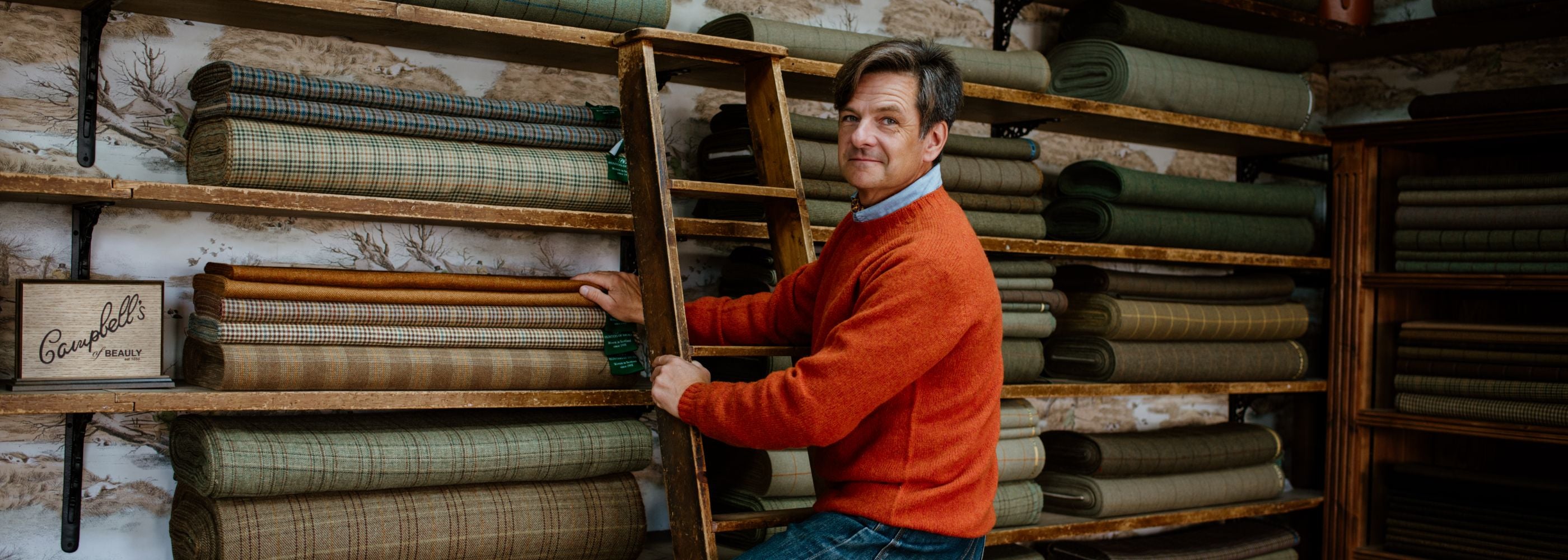 A man in an orange sweater sits on a wooden ladder in front of shelves stacked with neatly folded rolls of plaid and solid fabric in various shades of green and brown.