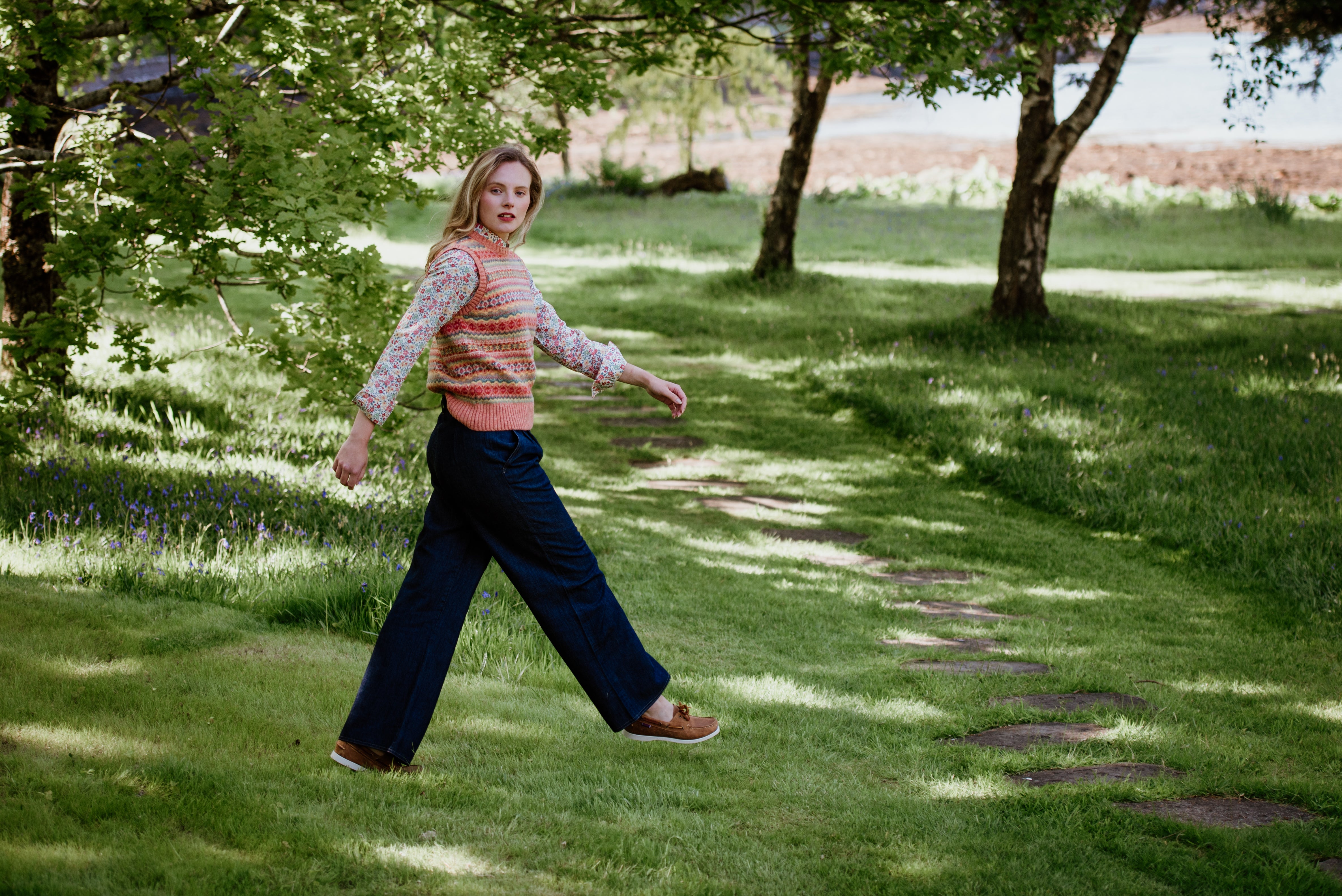 A woman with blonde hair wearing a colorful sweater vest and wide-leg jeans walks along a stone path in a grassy, tree-filled park, glancing back over her shoulder.