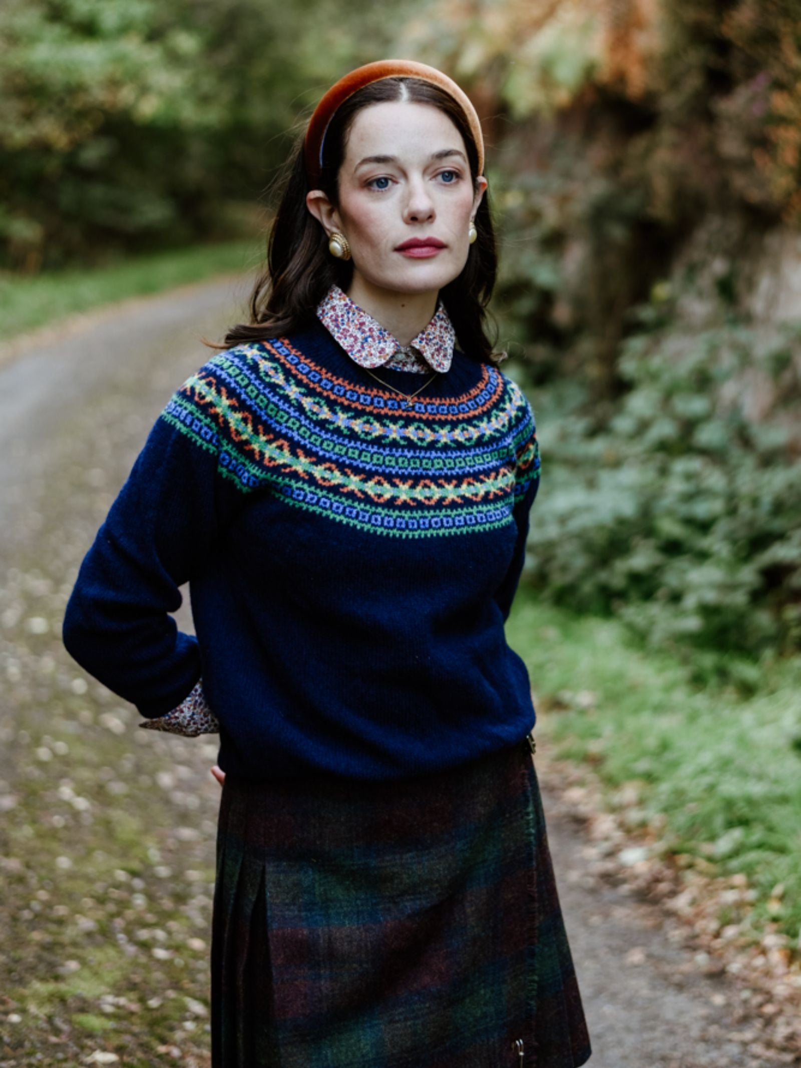 A woman with dark hair and a thoughtful expression stands on a tree-lined path wearing Campbell's of Beauly's Auld Stock Fairisle Crew Jumper, crafted from soft Geelong lambswool in Scotland, styled with a collared shirt, tartan skirt, and orange headband.
