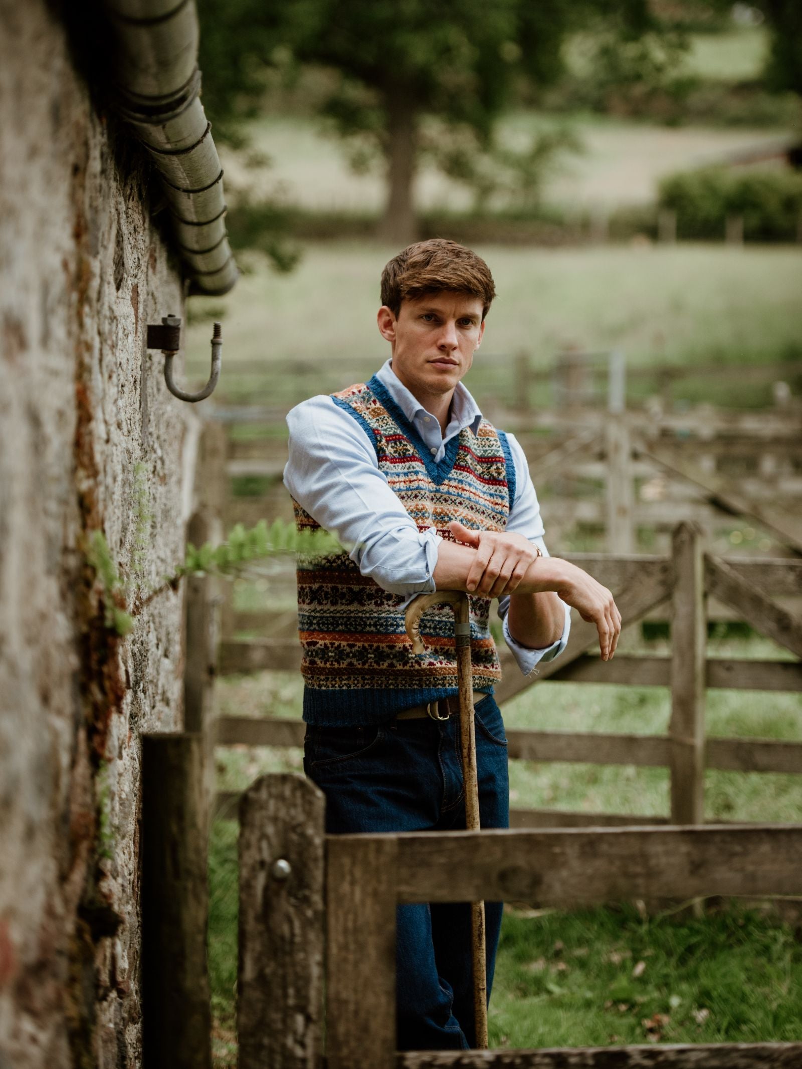 A man in a Campbell's of Beauly Fairisle Slipover and blue shirt leans on a stick by a stone wall and wooden fence in a rural outdoor setting with green fields behind him.