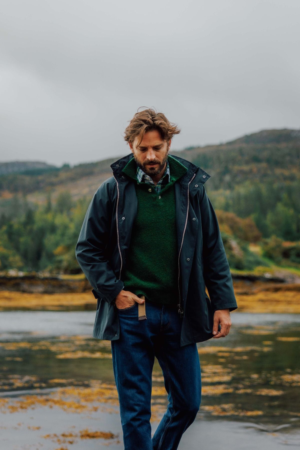 A man with wavy hair wearing a dark rain jacket, green sweater, plaid shirt, and jeans stands outdoors on a cloudy day, with hills and trees in the background.