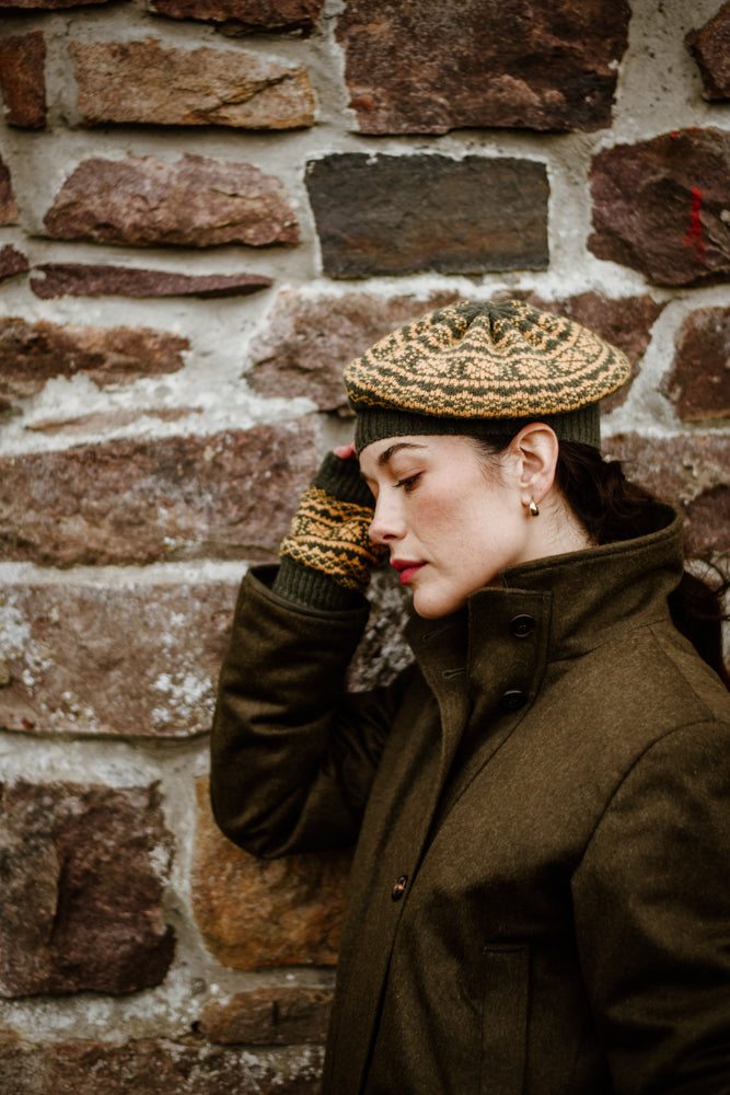 A woman in an olive coat, patterned knit hat, and matching gloves leans her head against a stone wall, eyes closed, with a calm and thoughtful expression.