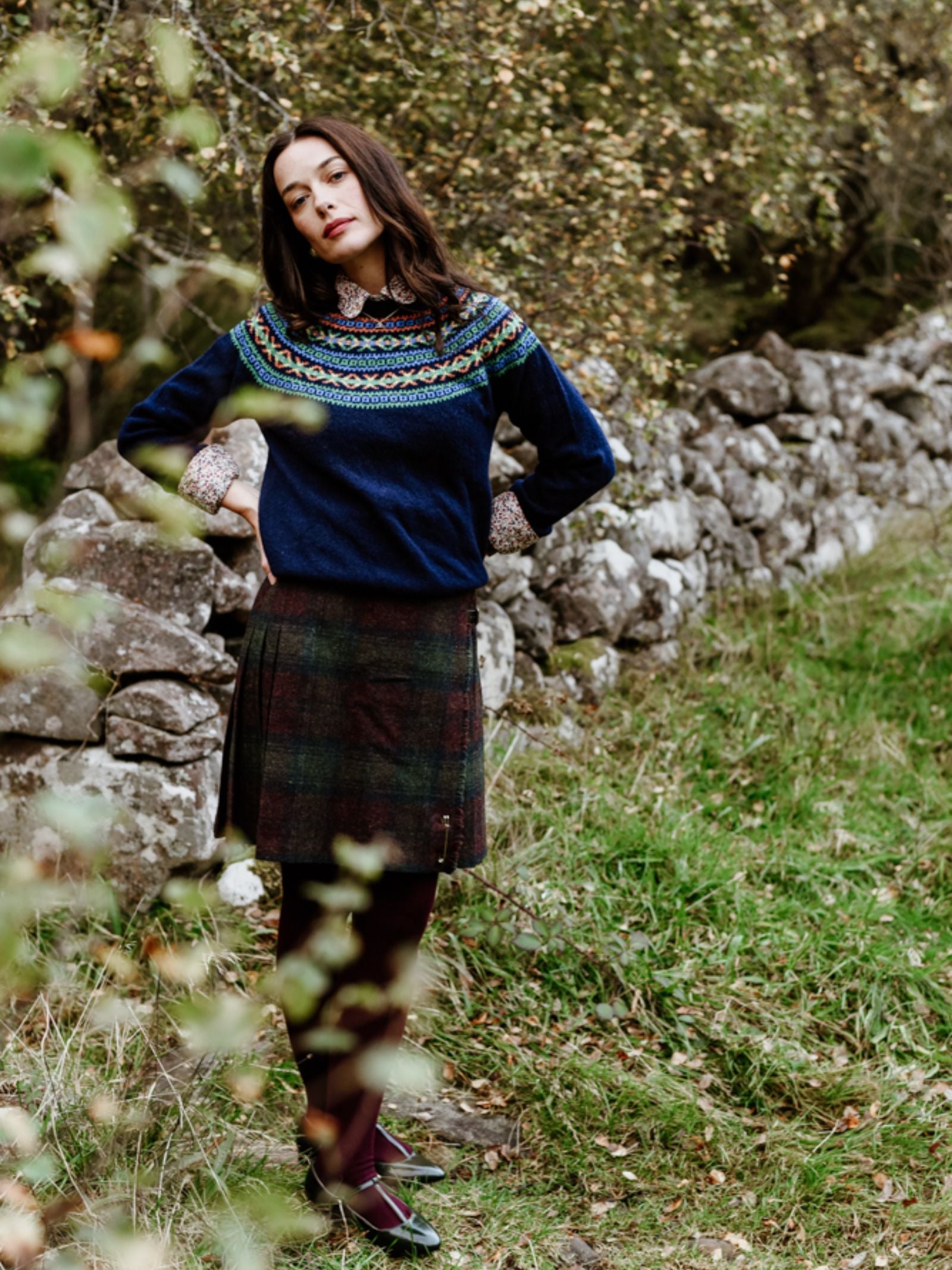 A woman stands outdoors by a stone wall, wearing Campbell's of Beauly's Auld Stock Fairisle Crew Jumper in blue, paired with a plaid skirt, dark tights, and black shoes amid greenery and overhanging branches.
