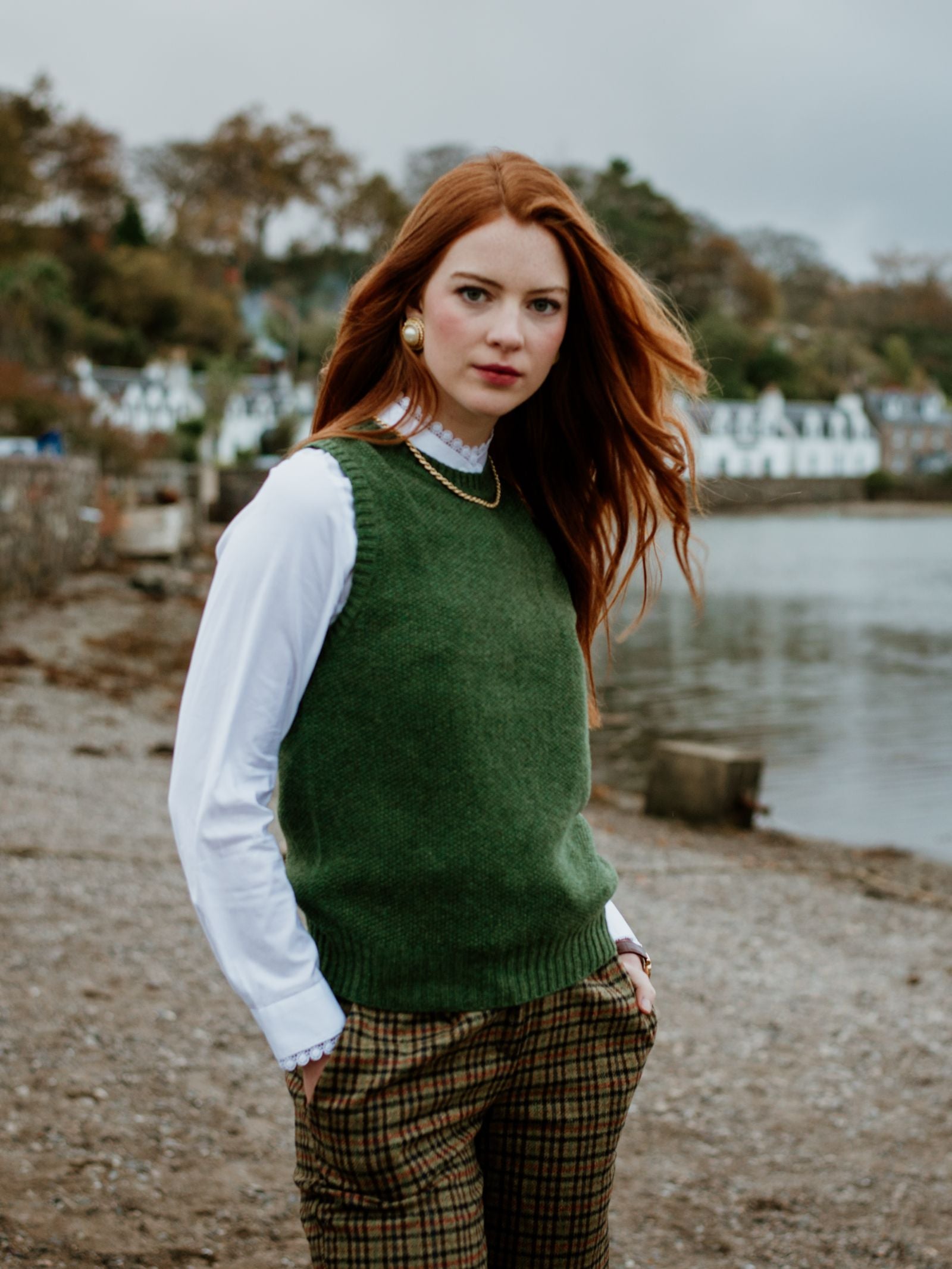 A woman with long red hair stands by a lakeshore, hands in pockets, wearing the Campbell's of Beauly Auld Stock Moss Stitch Crew Tank over a white blouse and plaid pants. Houses and trees appear in the background beneath a cloudy sky.
