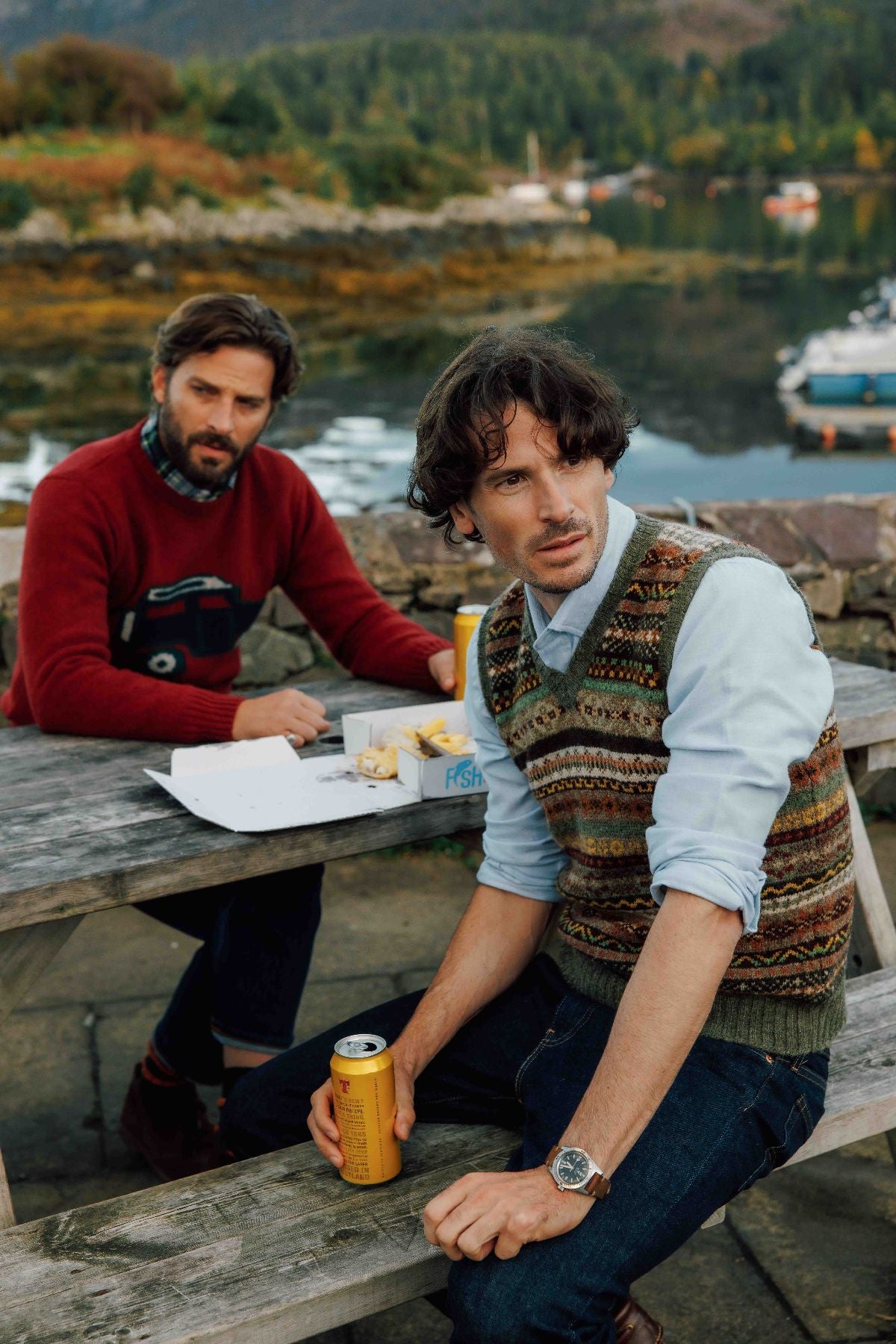 Two men sit at a wooden picnic table outdoors by a lake with hills and boats in the background. One man in a patterned sweater vest holds a yellow can, while the other, in a red sweater, sits further back with food.