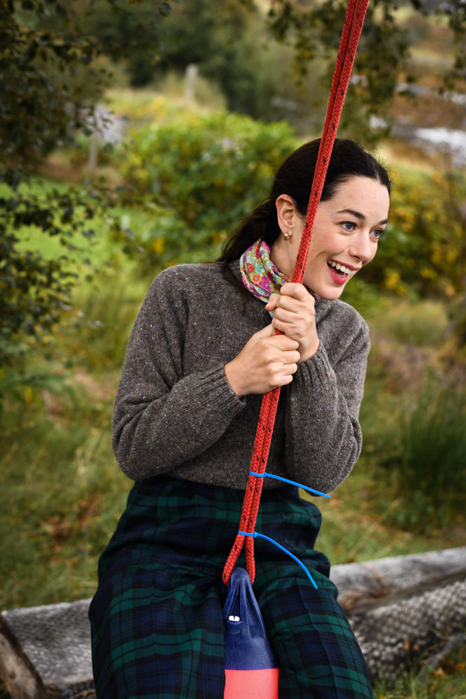 A young woman with dark hair, wearing a brown sweater and plaid skirt, smiles excitedly while sitting on a red rope swing outdoors, surrounded by greenery.