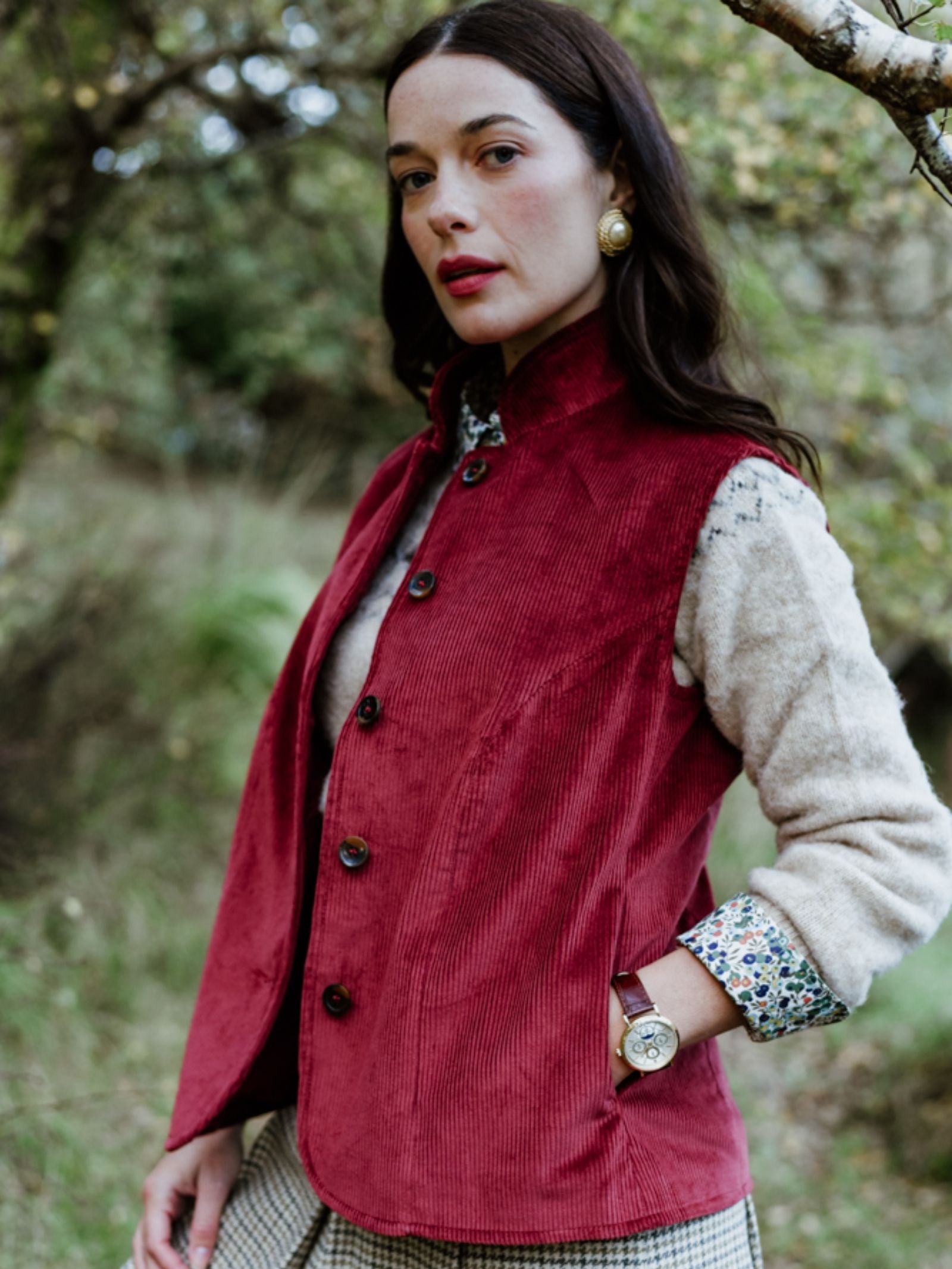 A woman with long dark hair stands outdoors among trees, wearing Campbell's of Beauly's Auld Stock Corduroy Gilet in red over a beige sweater, plaid skirt, gold earrings, and a watch, exuding country elegance.