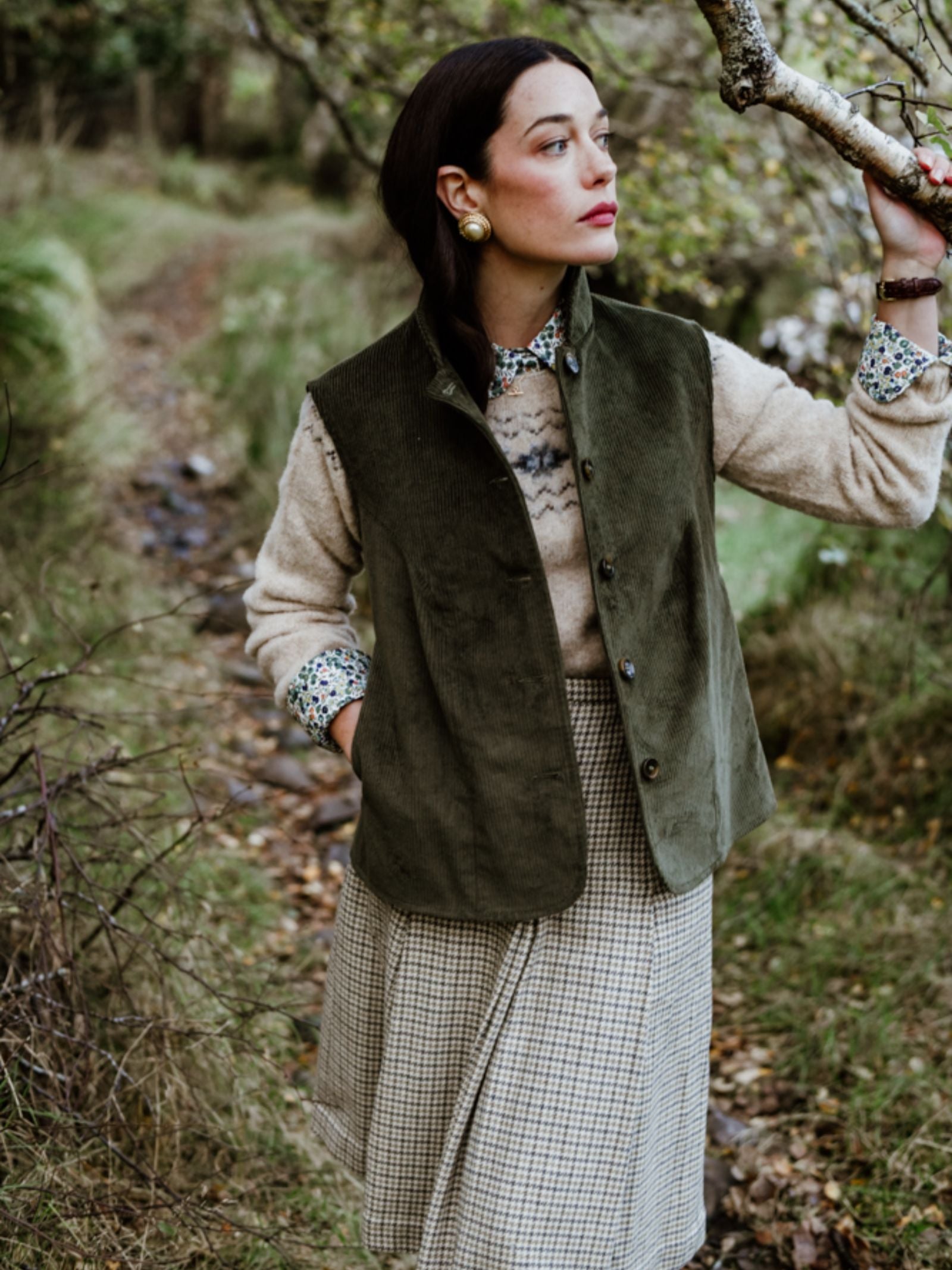 In the countryside, a woman embodies country elegance in Campbell’s of Beauly’s Auld Stock Corduroy Gilet, paired with a beige patterned sweater, floral shirt, and plaid skirt as she stands by a tree on a leaf-strewn path.