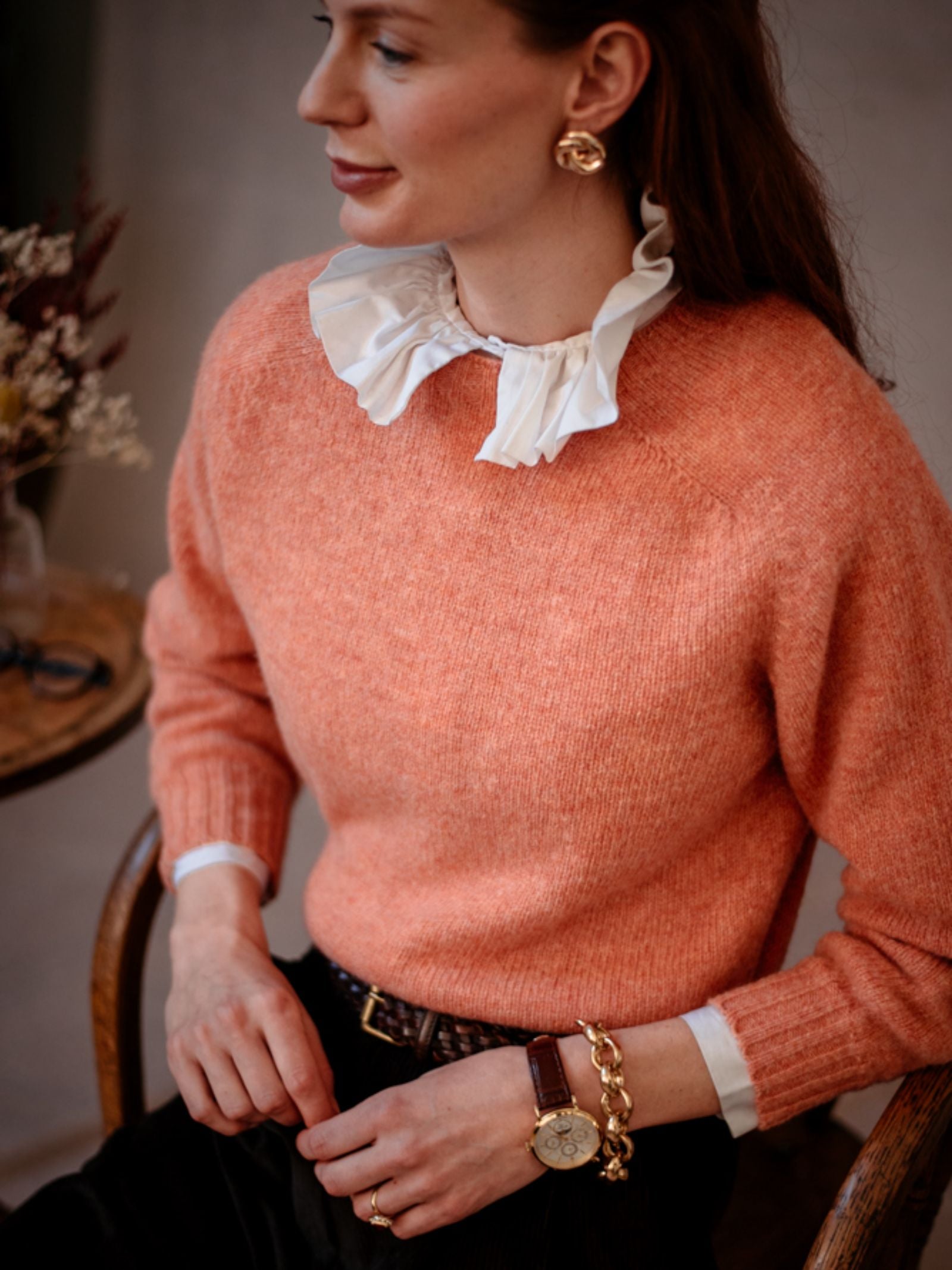 A woman with long brown hair sits on a chair, turned slightly to the side, wearing a peach Auld Stock Cropped Shetland Jumper by Campbell's of Beauly over a white ruffled-collar shirt, accessorized with gold earrings, a gold bracelet, and a watch.