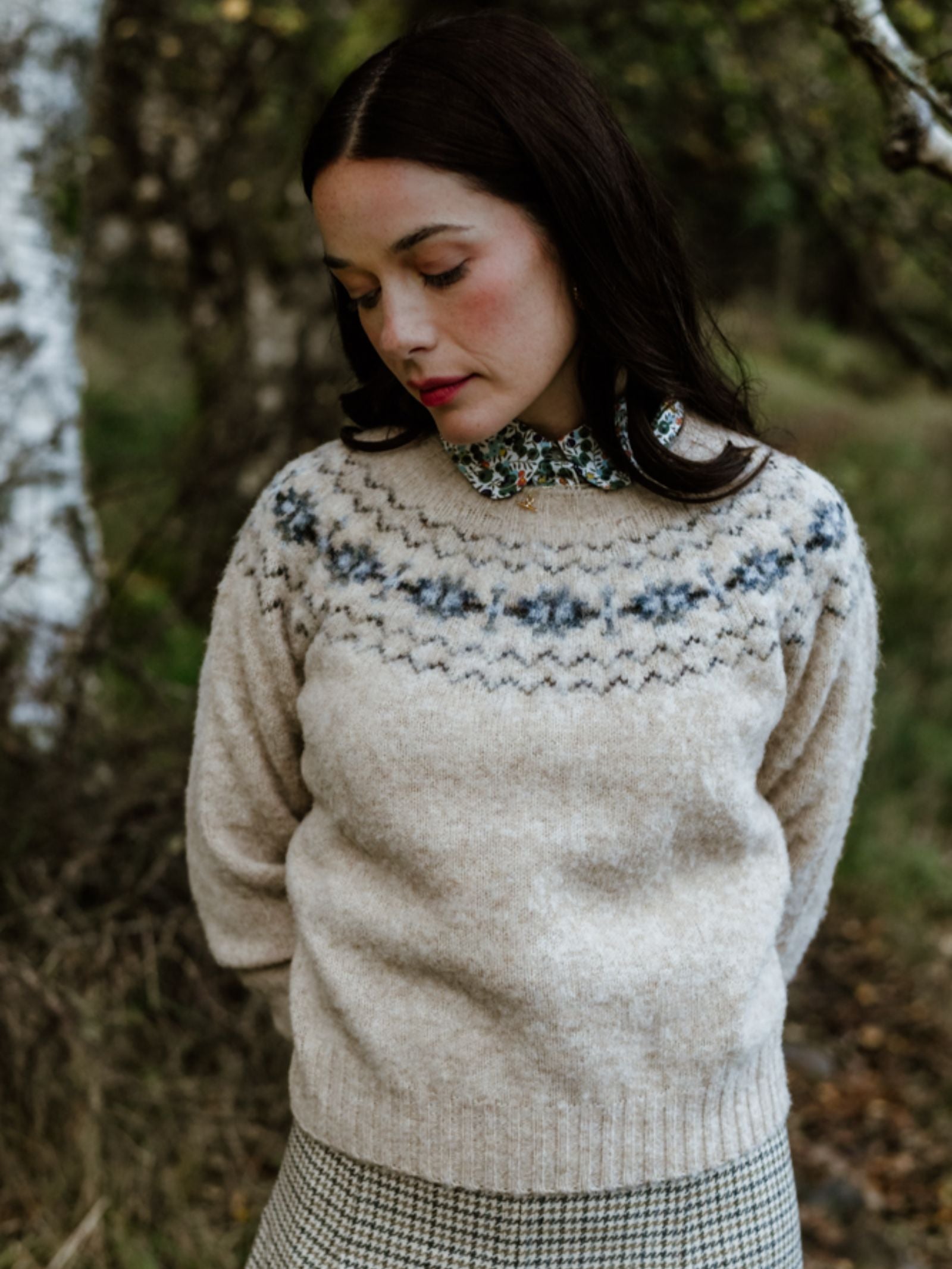 A woman with long dark hair stands outdoors among trees, wearing the Shaggy Cropped Fairisle Crew Jumper by Campbell's of Beauly over a floral-collared shirt and checkered skirt, gazing down with a serene expression.