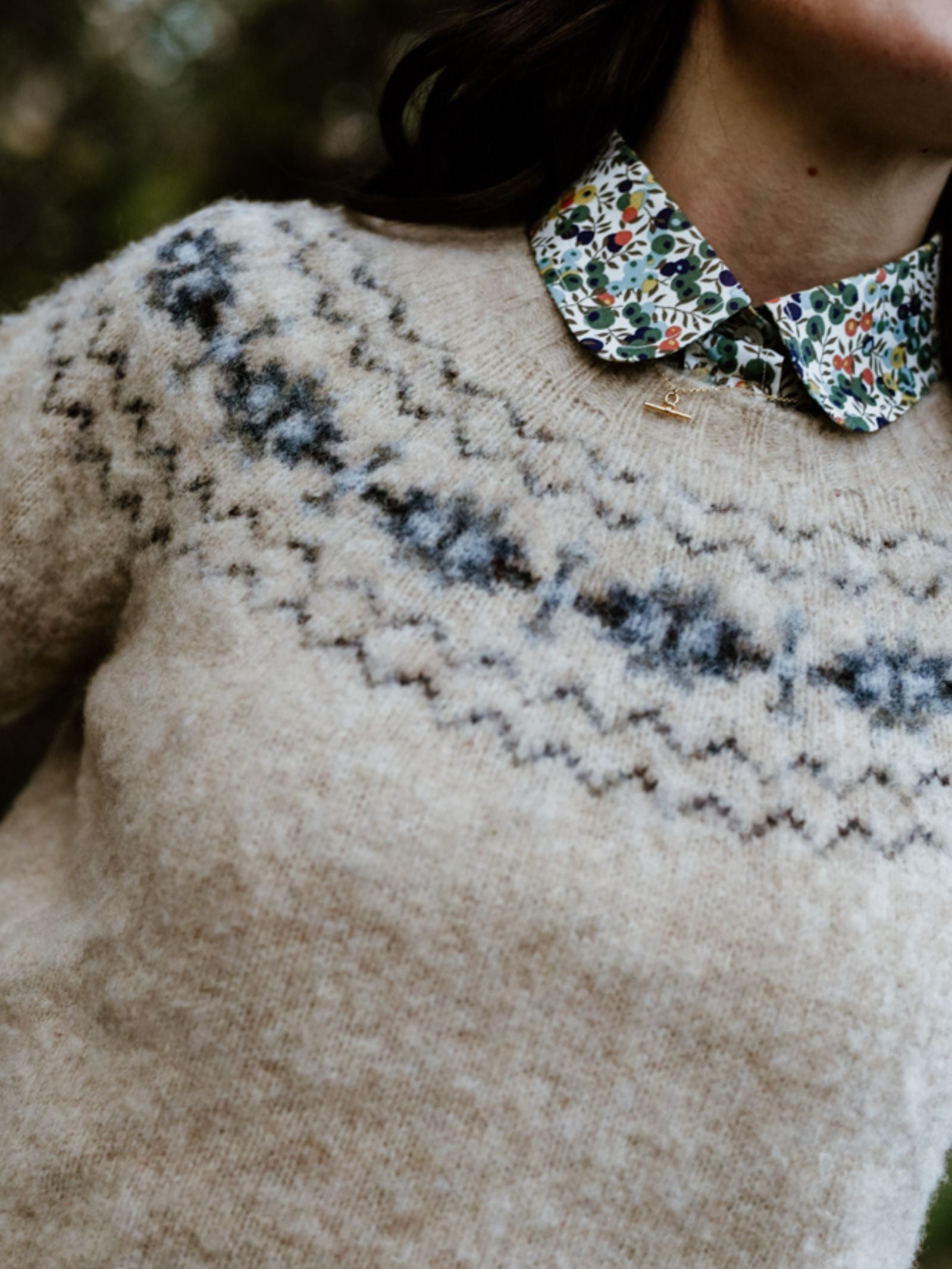 A person outdoors wears the Campbell's of Beauly Shaggy Cropped Fairisle Crew Jumper in beige with blue and gray zigzags, layered over a floral collared shirt. Only their lower face, neck, and upper torso are visible.