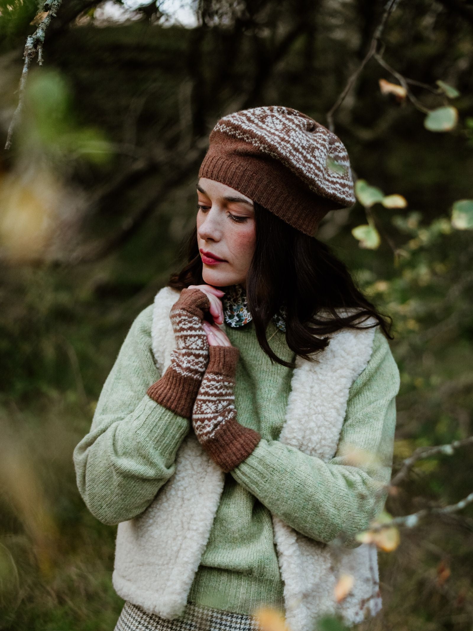 A woman in a green sweater and cream vest stands outdoors, wearing Campbell's of Beauly Auld Stock 2-Col Half Finger Gloves and a brown fairisle beret, looking thoughtfully with dark hair in a wooded area.